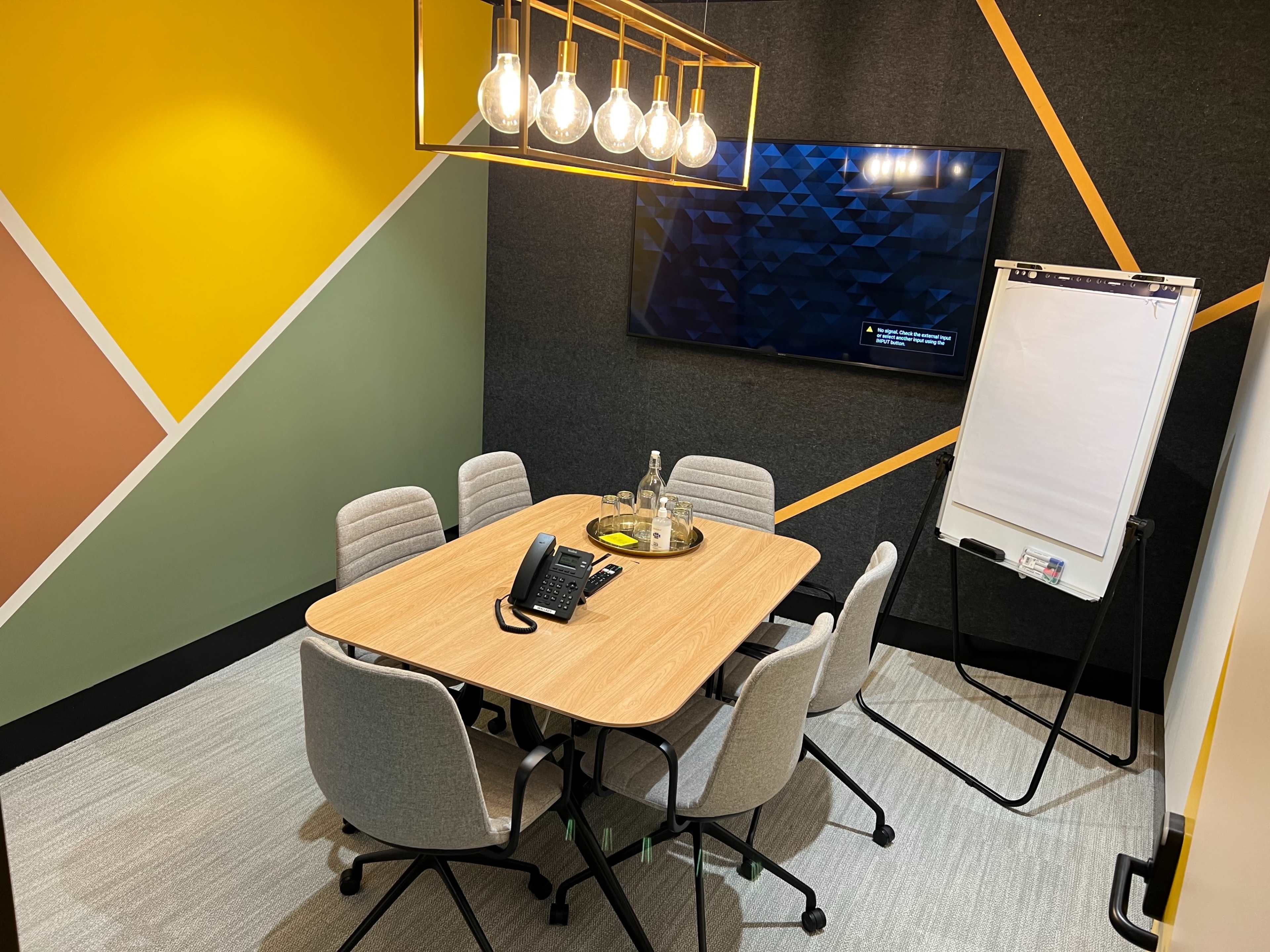 The image shows a small meeting room with a wooden table, six chairs, a large screen on the wall, and a whiteboard on a stand.