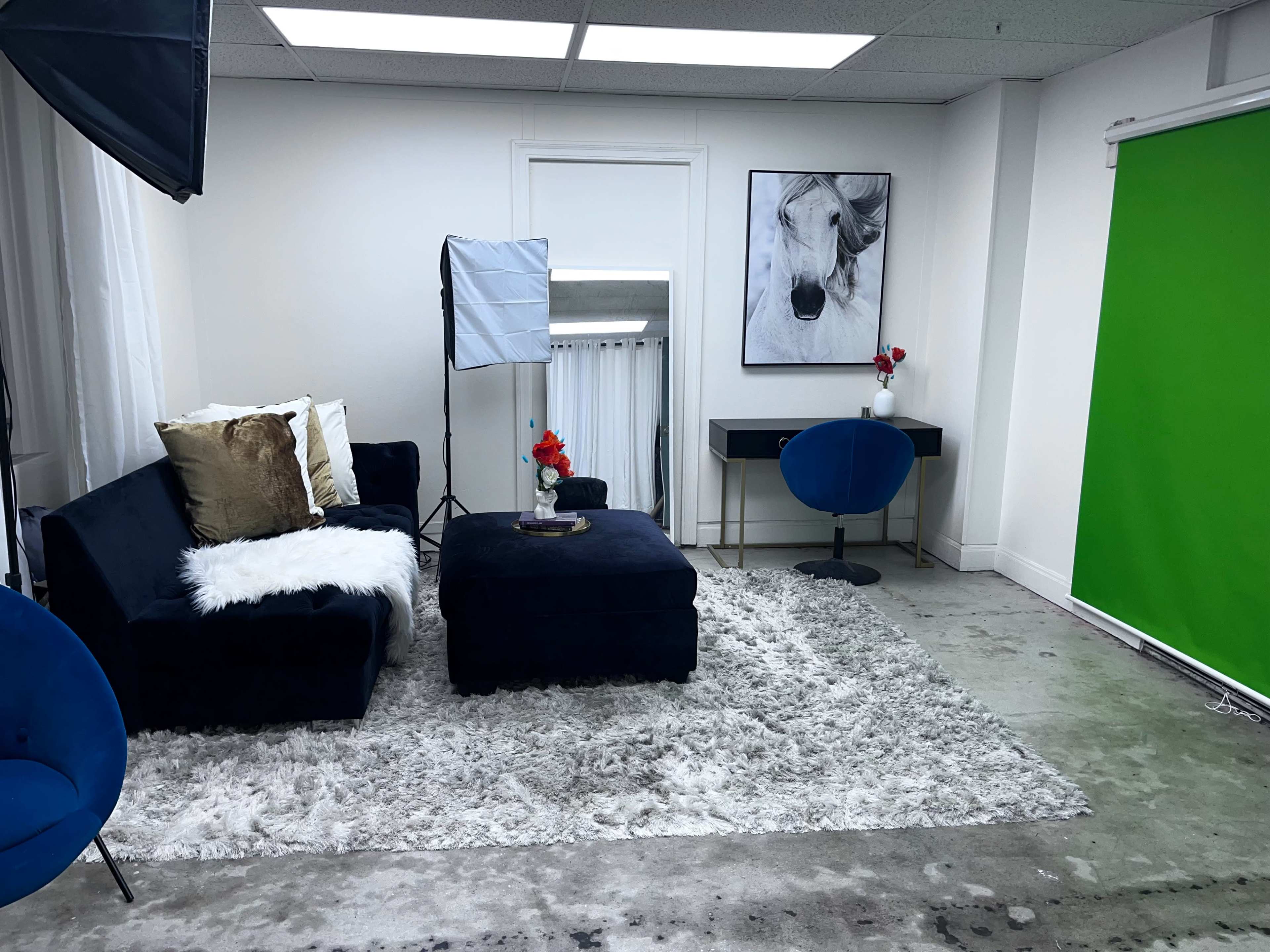 A styled interior space featuring a dark blue sofa, an ottoman, a decorative rug, and a green backdrop, along with a chair and a desk.