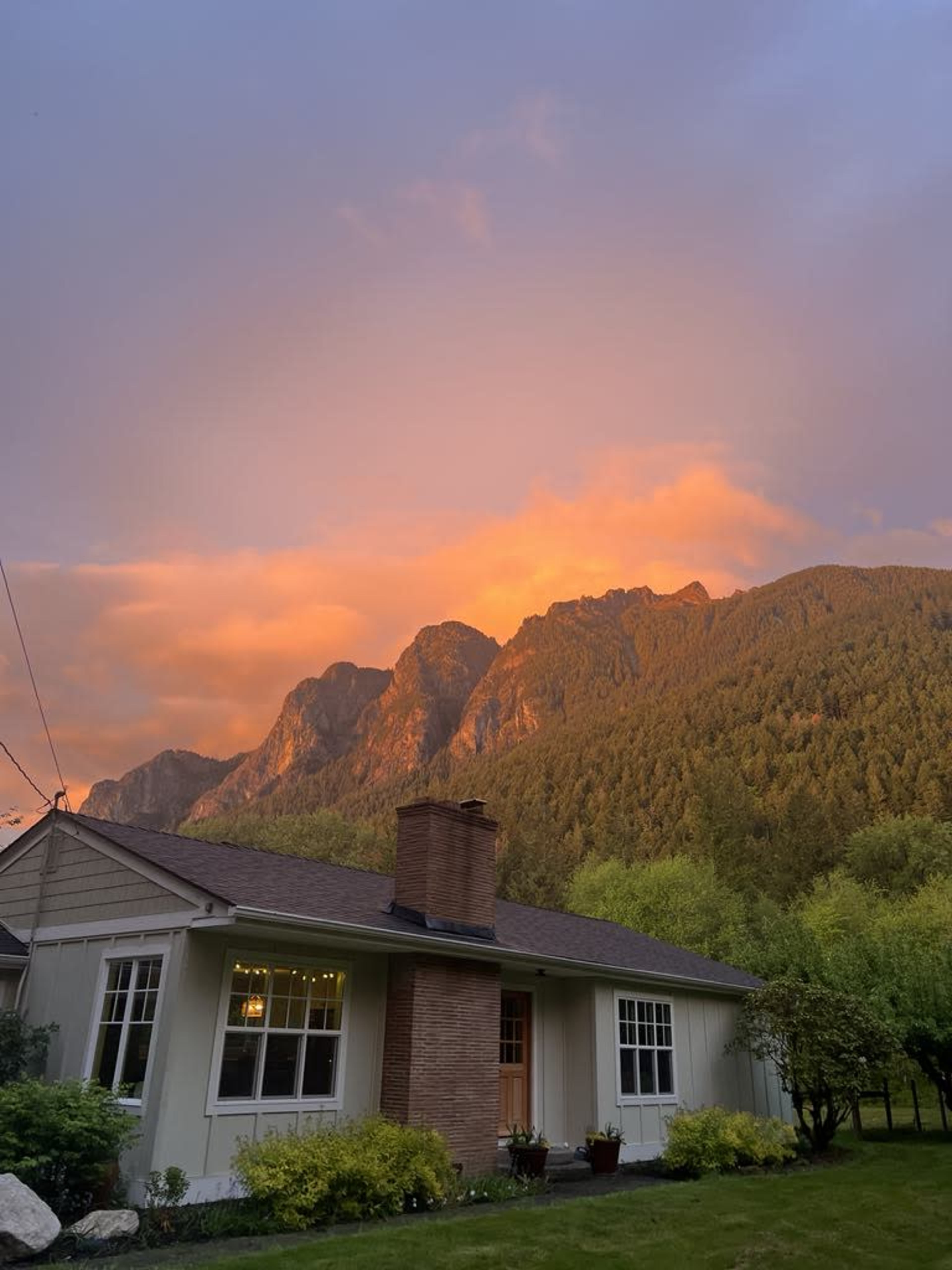 A house with a chimney is set against a backdrop of mountains illuminated by a vibrant sunset.
