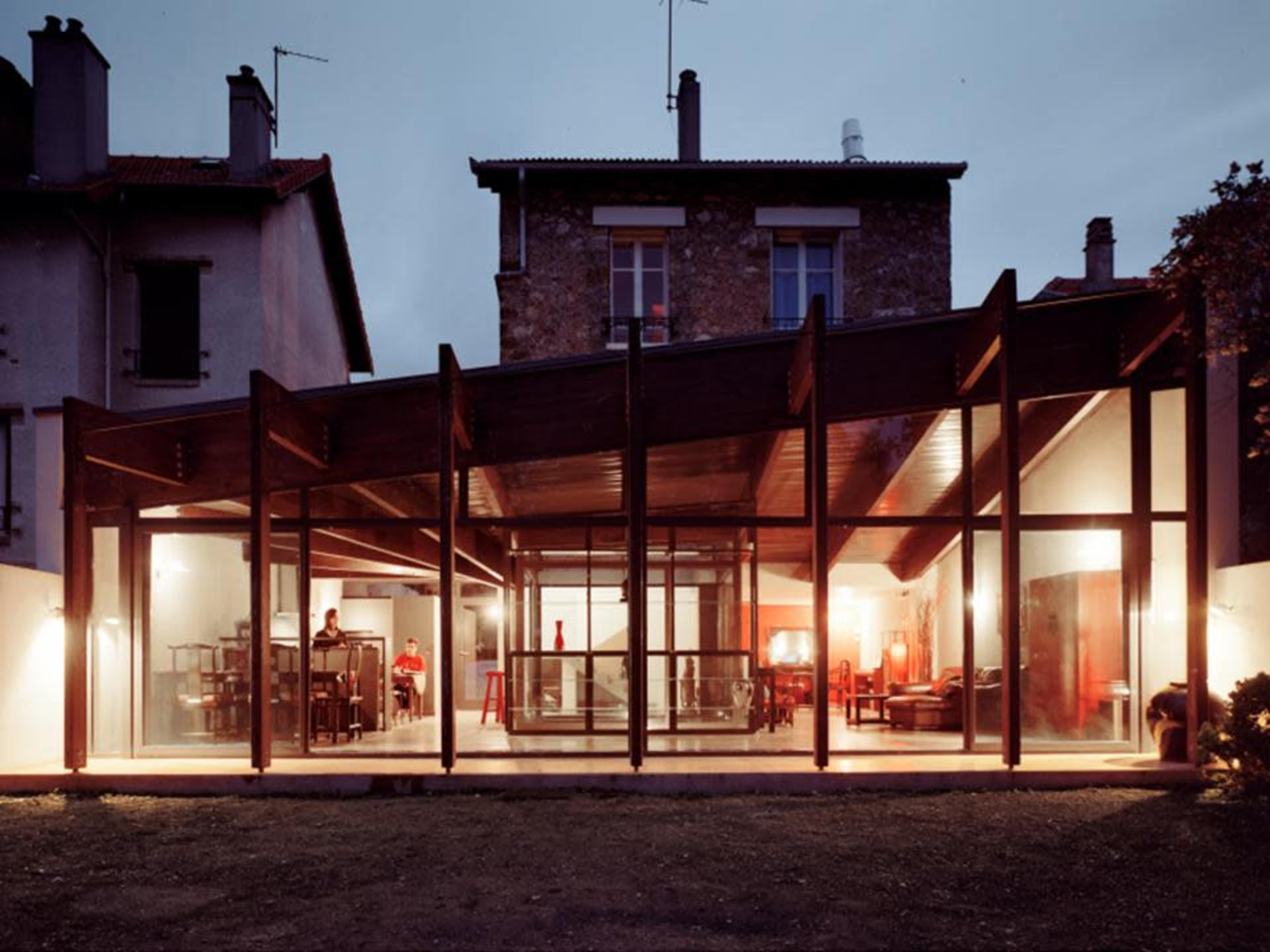 A modern house with a glass facade and wooden structure is illuminated at dusk, showcasing an open interior space.