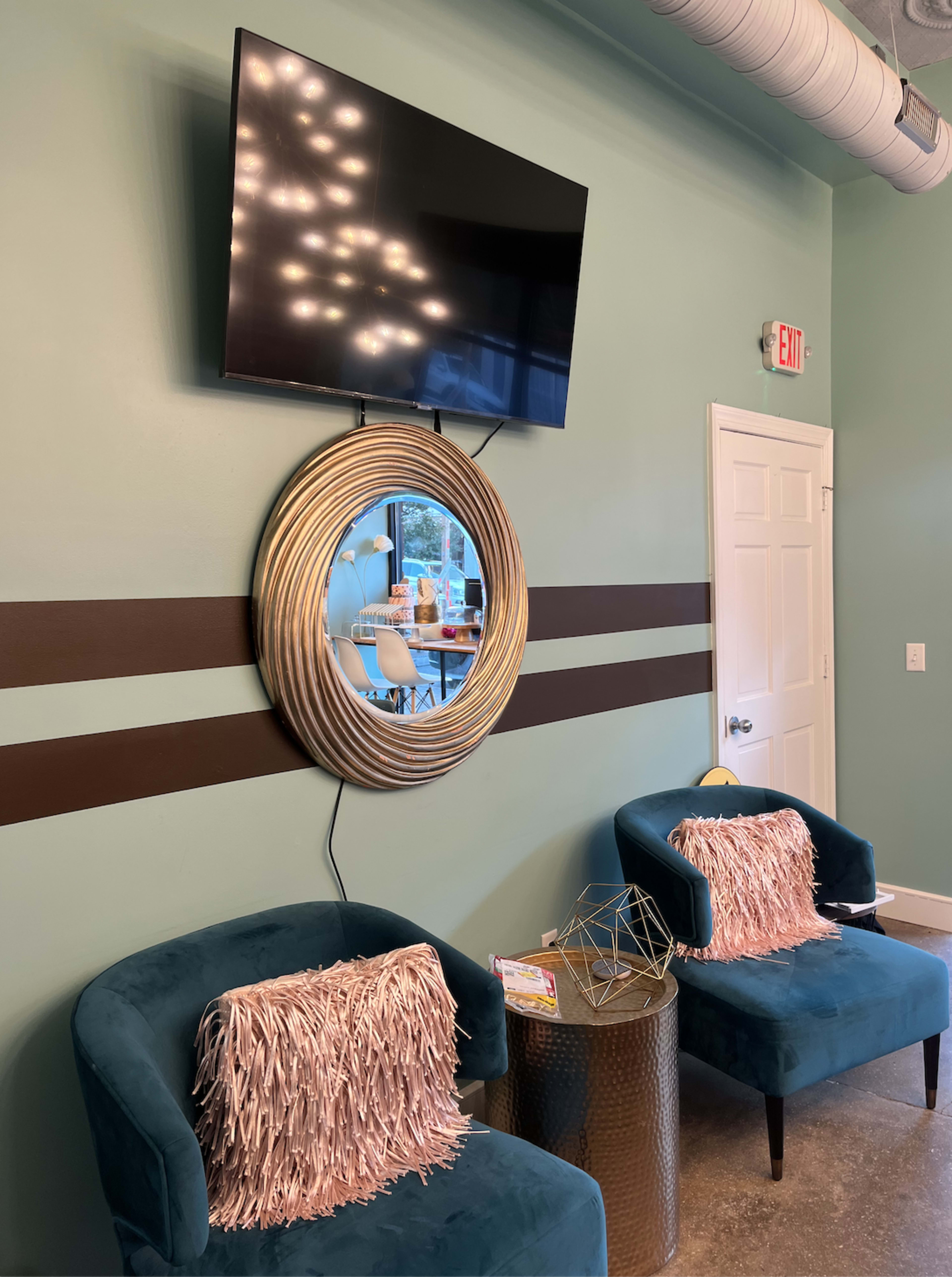 A modern waiting area with two teal chairs, a round mirror with a metallic frame, a mounted TV, and striped wall decor.