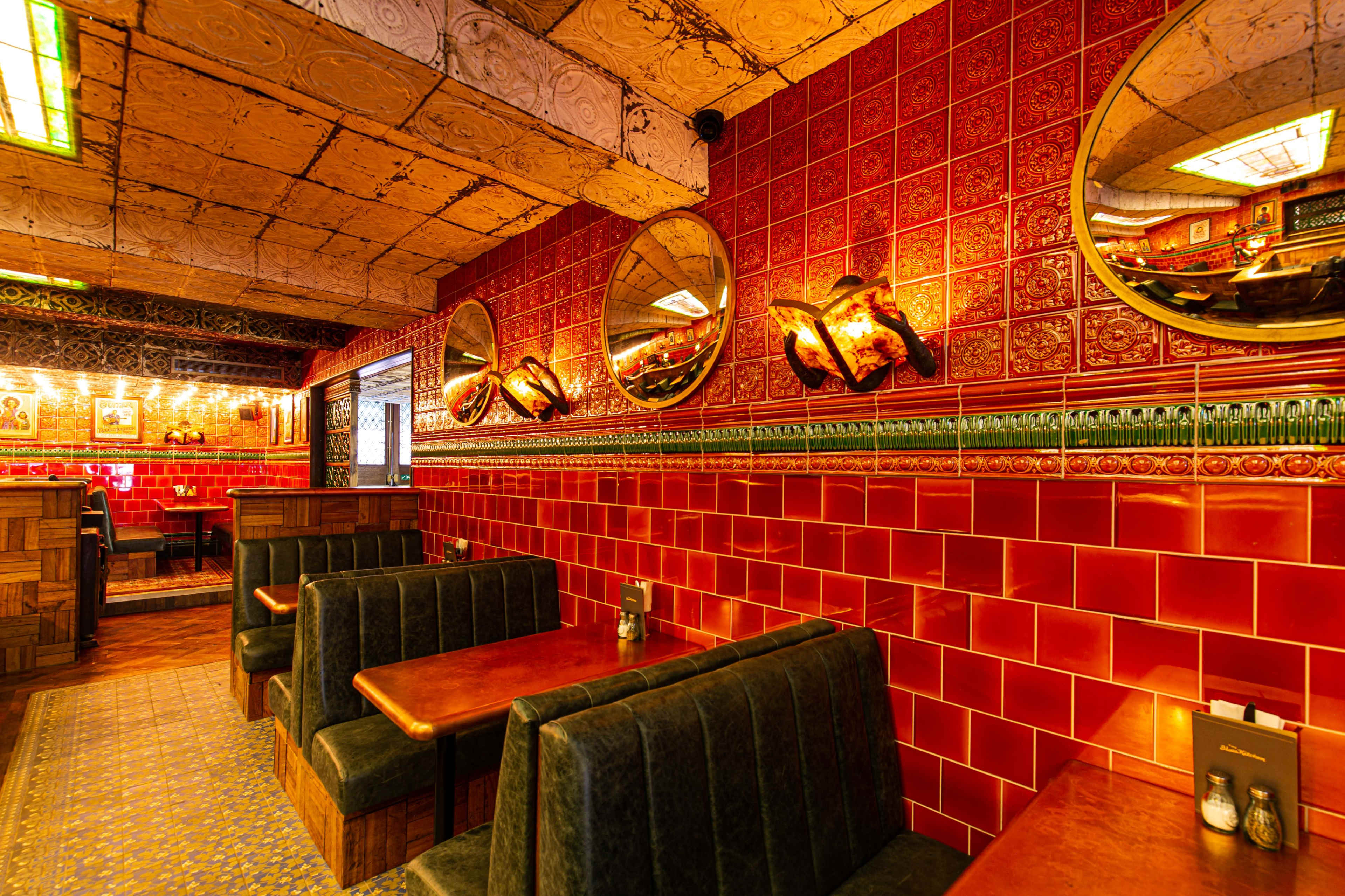 The interior of a restaurant features red tiled walls, wooden tables, and mirrors reflecting the warm lighting.