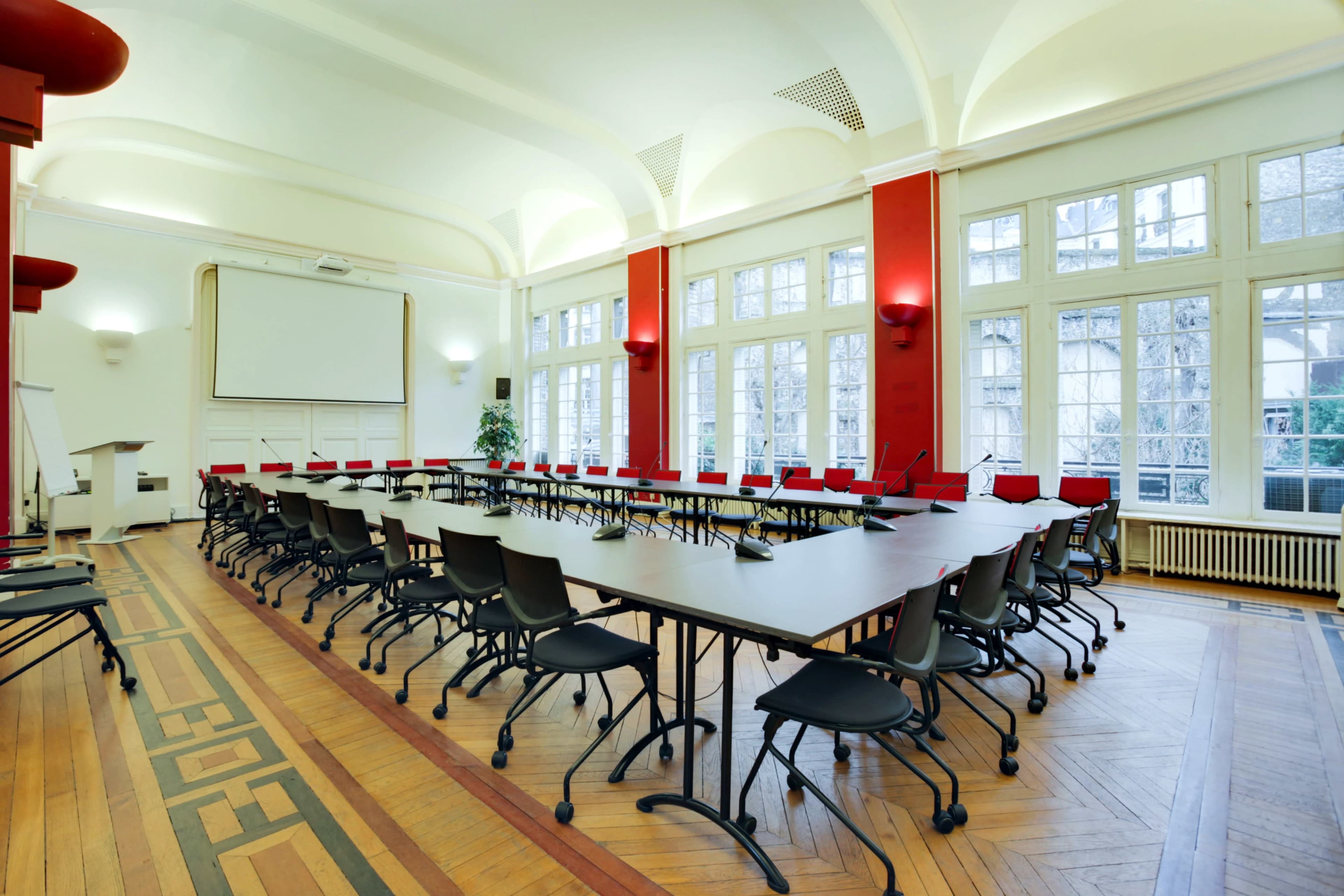 A large, empty conference room features several tables arranged in a U-shape surrounded by black chairs, with large windows letting in natural light.