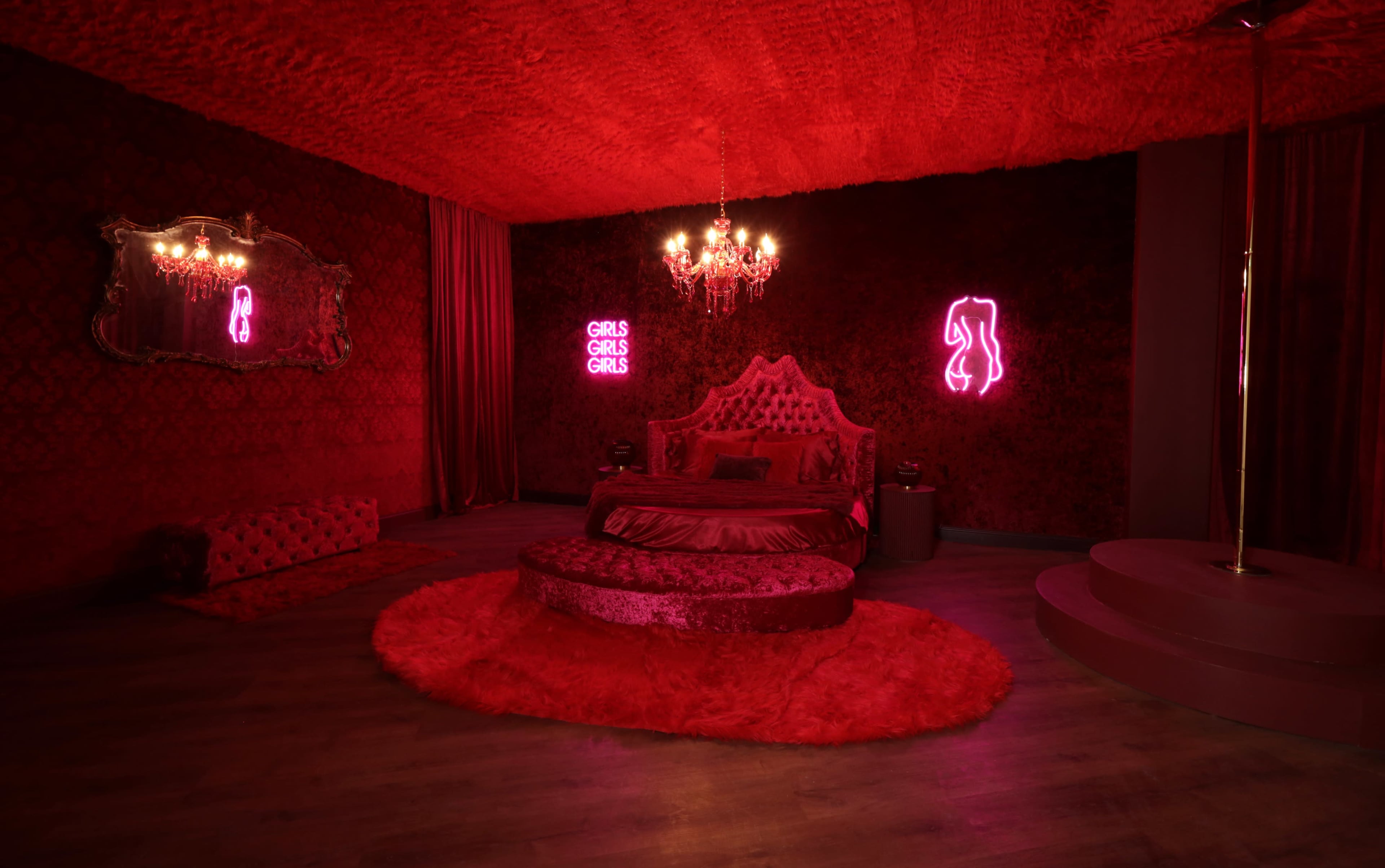 The scene showcases a dimly lit room featuring a plush, circular bed with a red velvet headboard, neon signs, and a pole, all set against richly textured red walls.