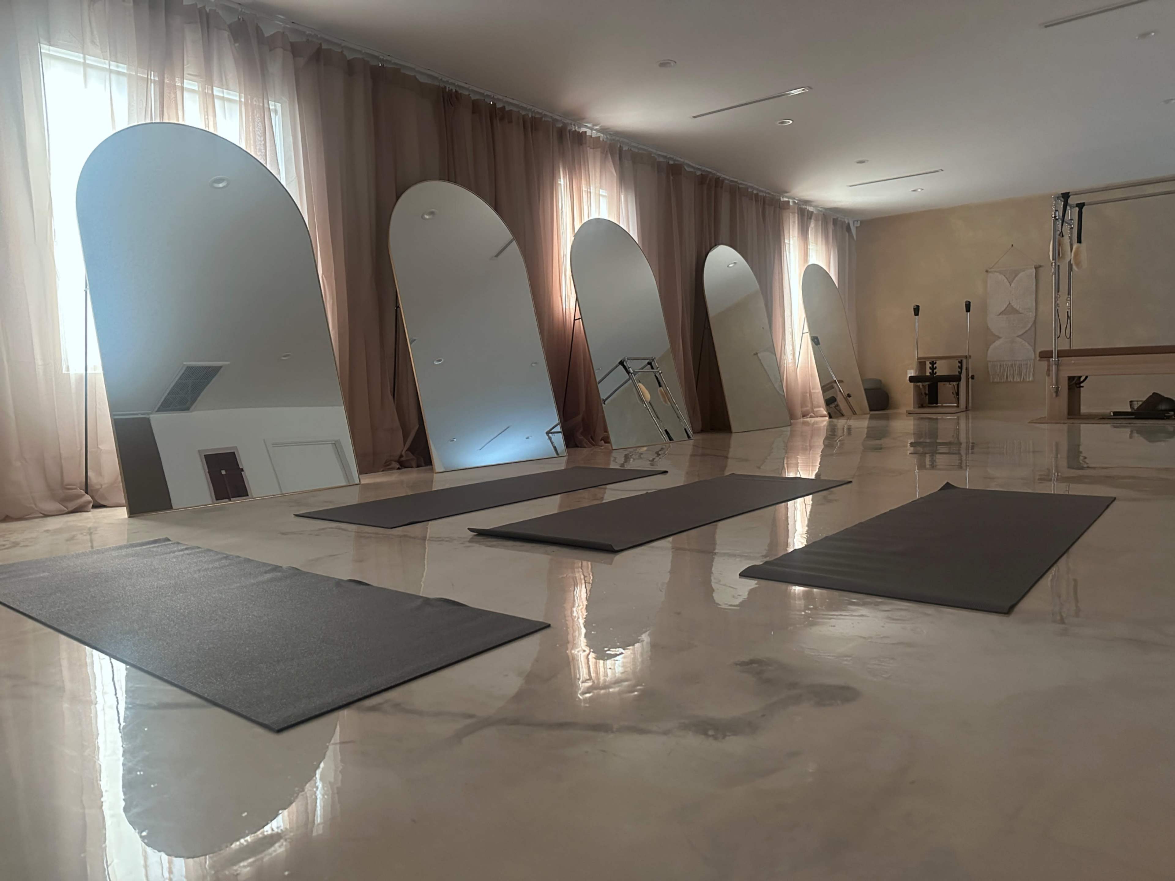 The image shows a spacious room with polished floors, featuring several yoga mats and large, arched mirrors along the walls.