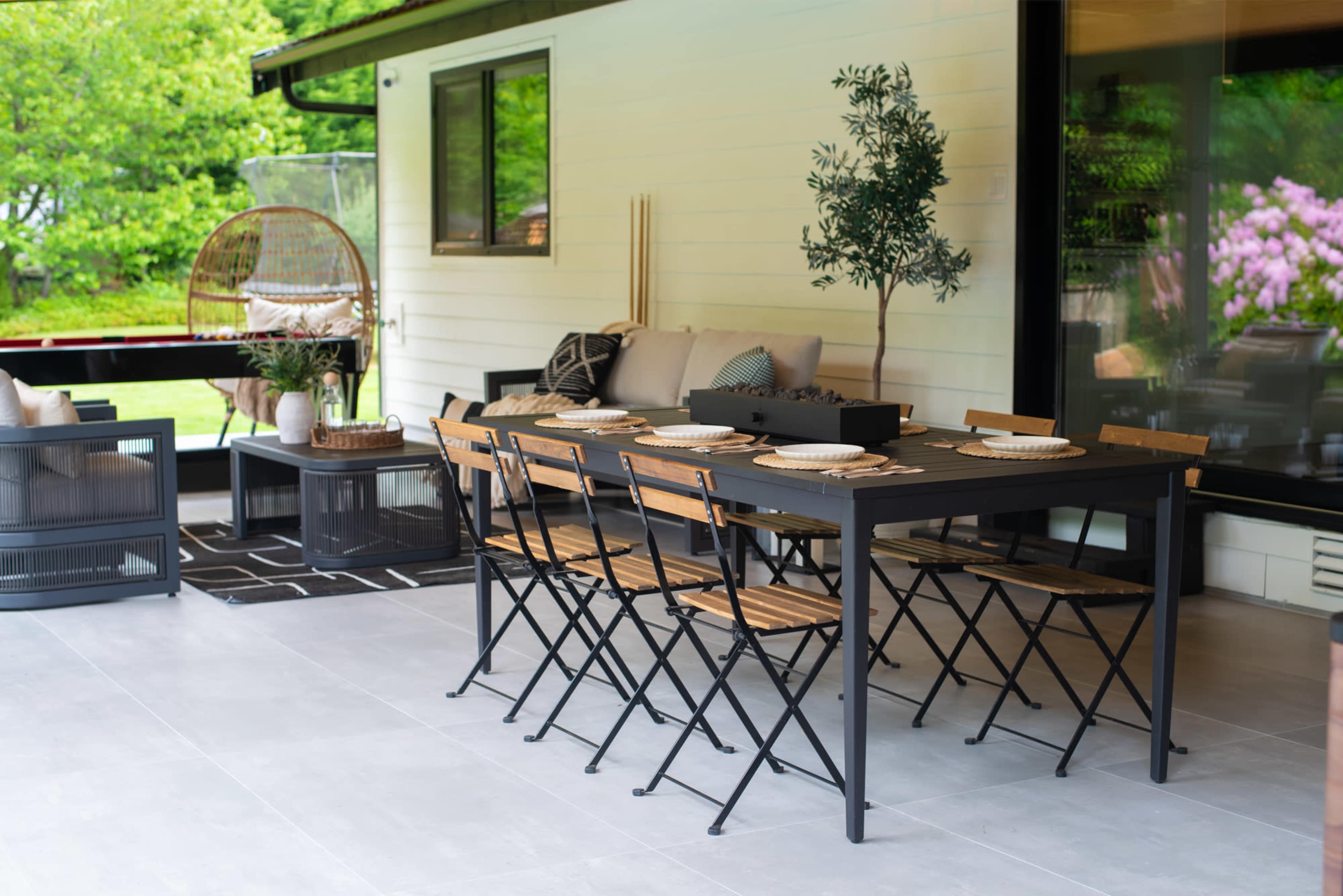 A modern outdoor dining area features a rectangular black table surrounded by wooden chairs, with a cozy seating arrangement and greenery in the background.