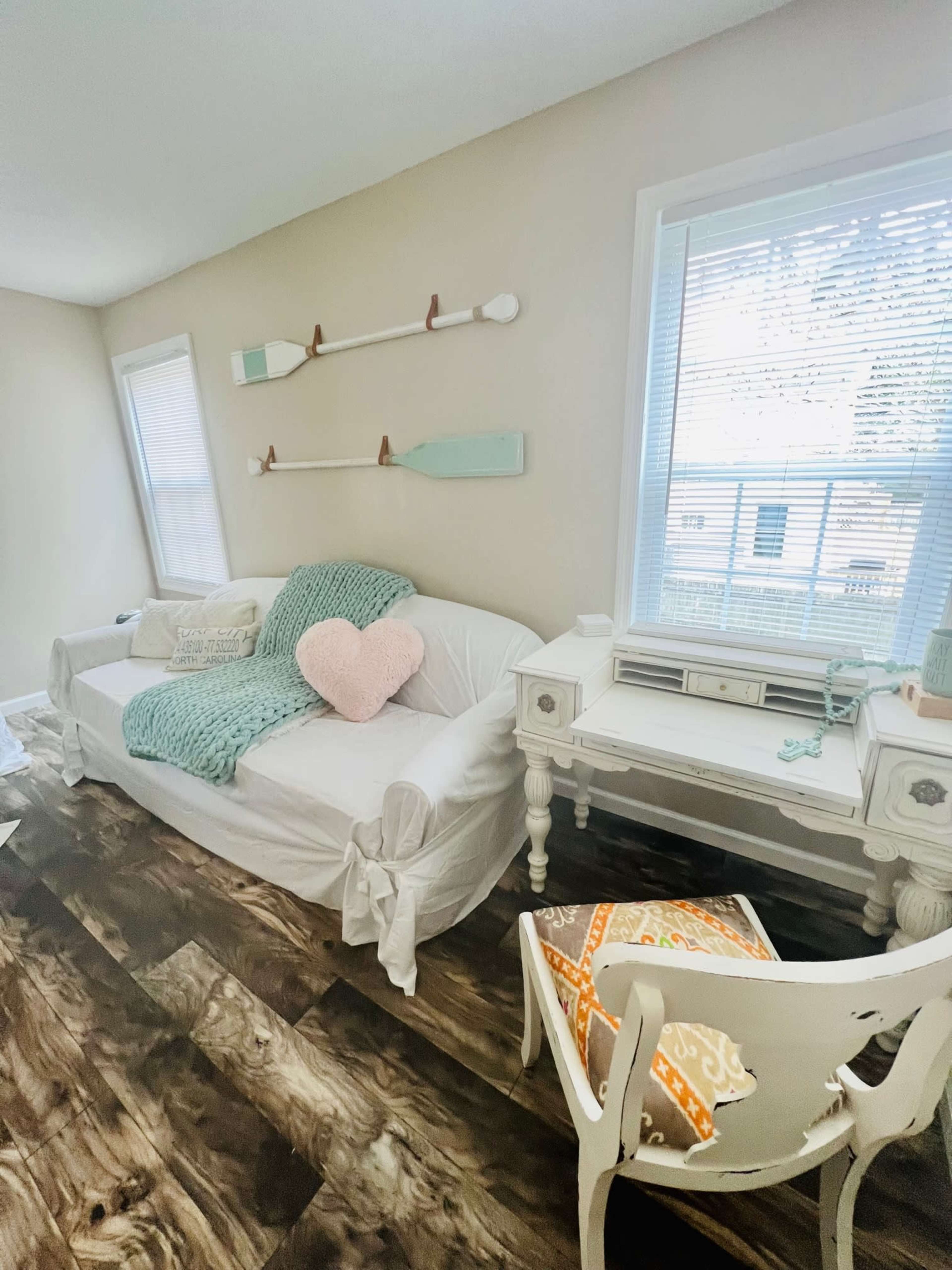 A bright living room featuring a white couch with a light green throw, a decorative heart pillow, and a desk beside a window with wooden flooring.