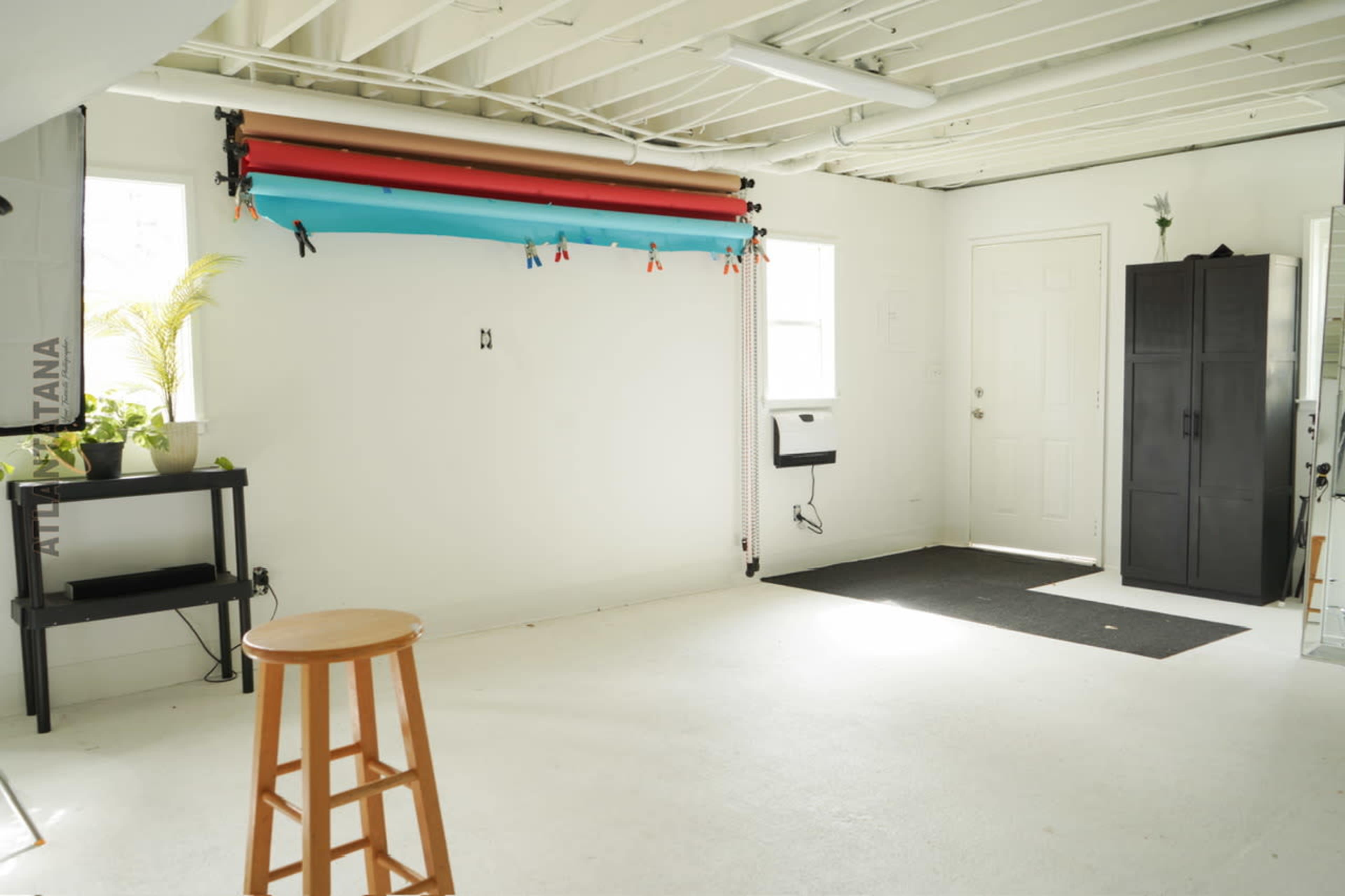 The image shows a spacious, well-lit room with white walls, light flooring, a black storage cabinet, a small table, and a backdrop setup for photography.