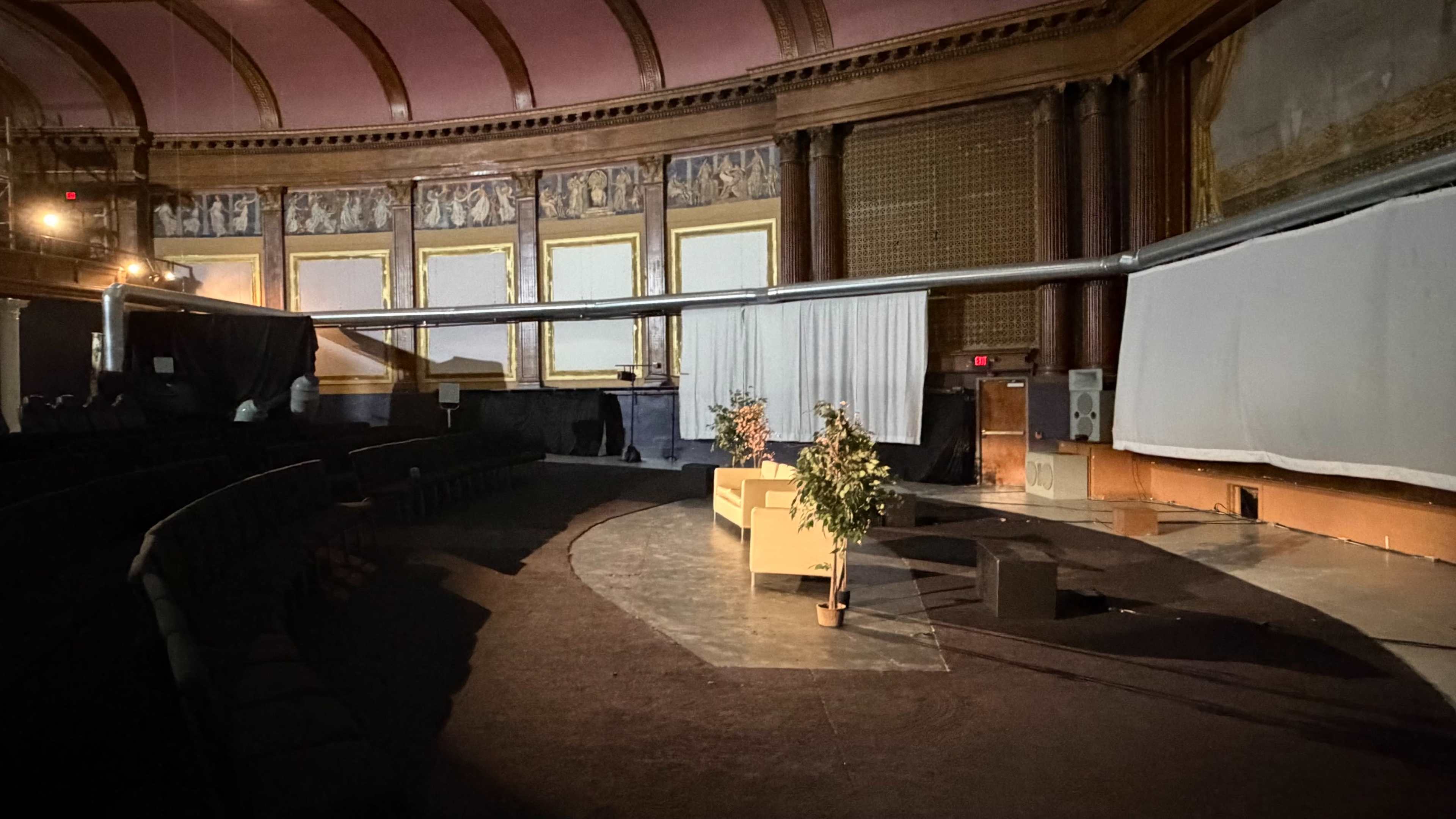 The image shows an empty theater with a curved seating arrangement, a long pipe extending across the stage, and a set of plants near a white backdrop.