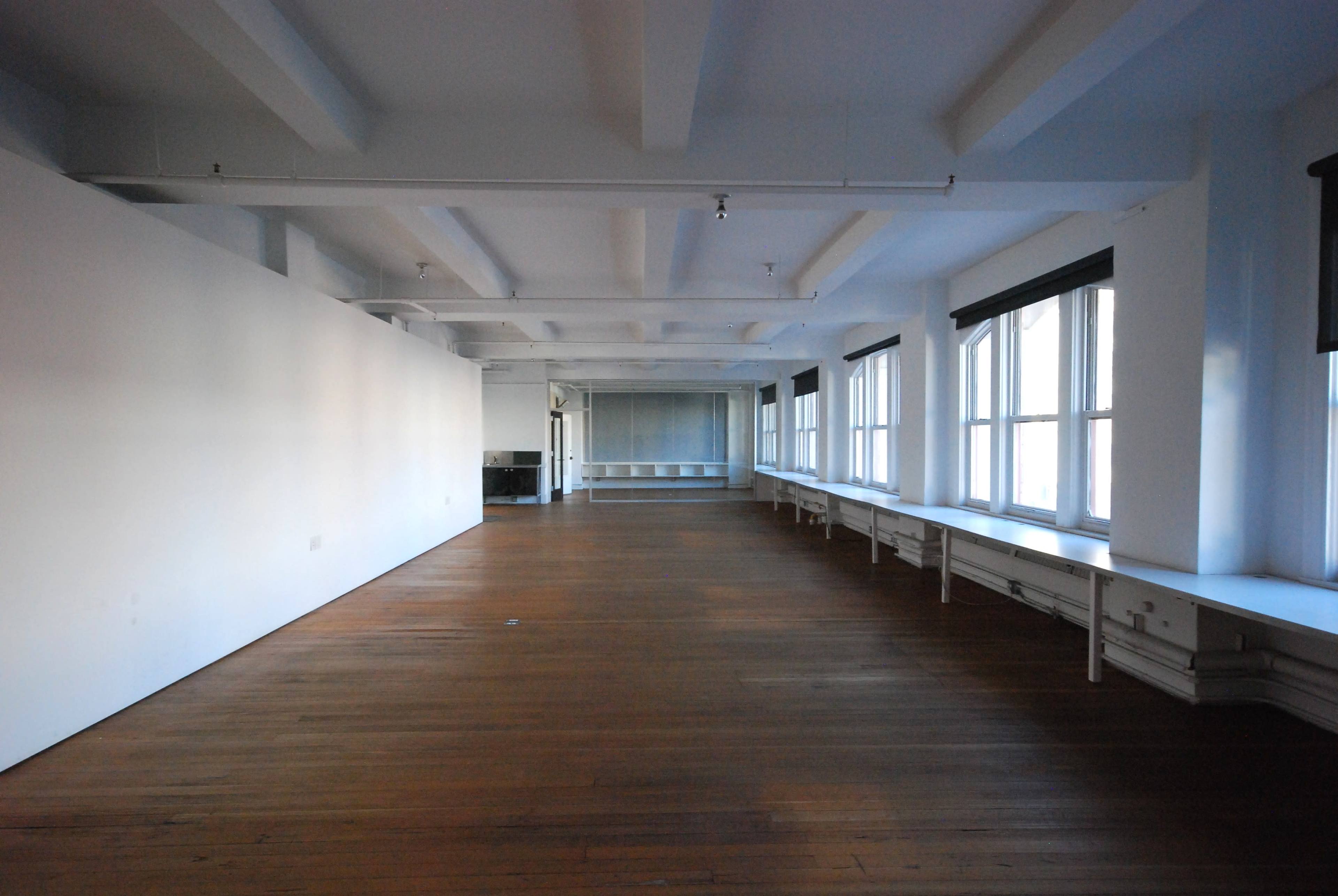 The image shows a spacious, empty room with wooden floors, large windows, and white walls.