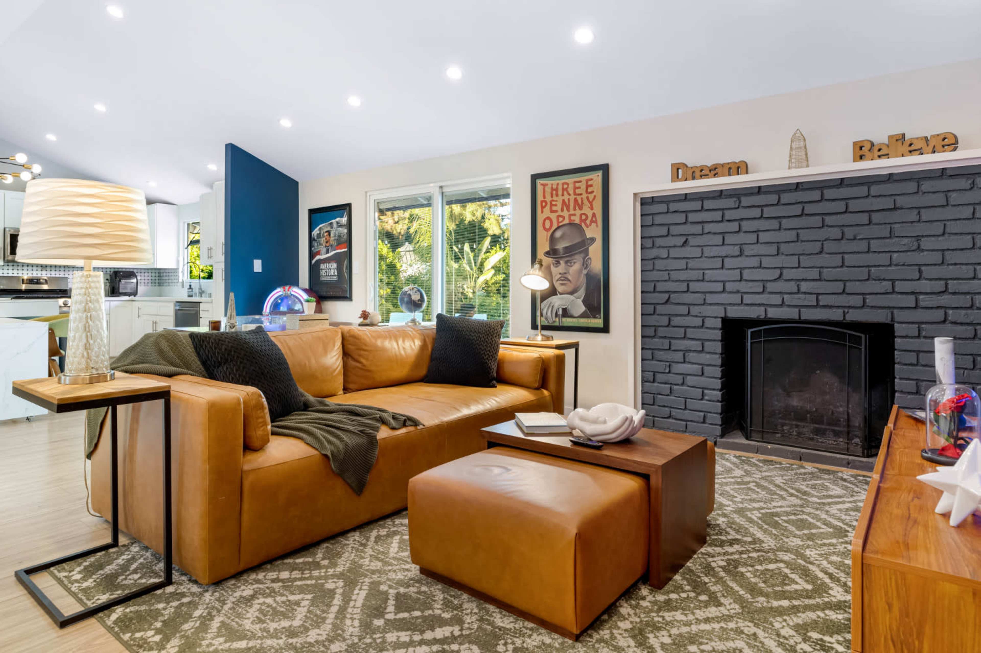 The image shows a modern living room with a brown leather couch, a patterned area rug, and a black brick fireplace, complemented by framed wall art and large windows revealing greenery outside.