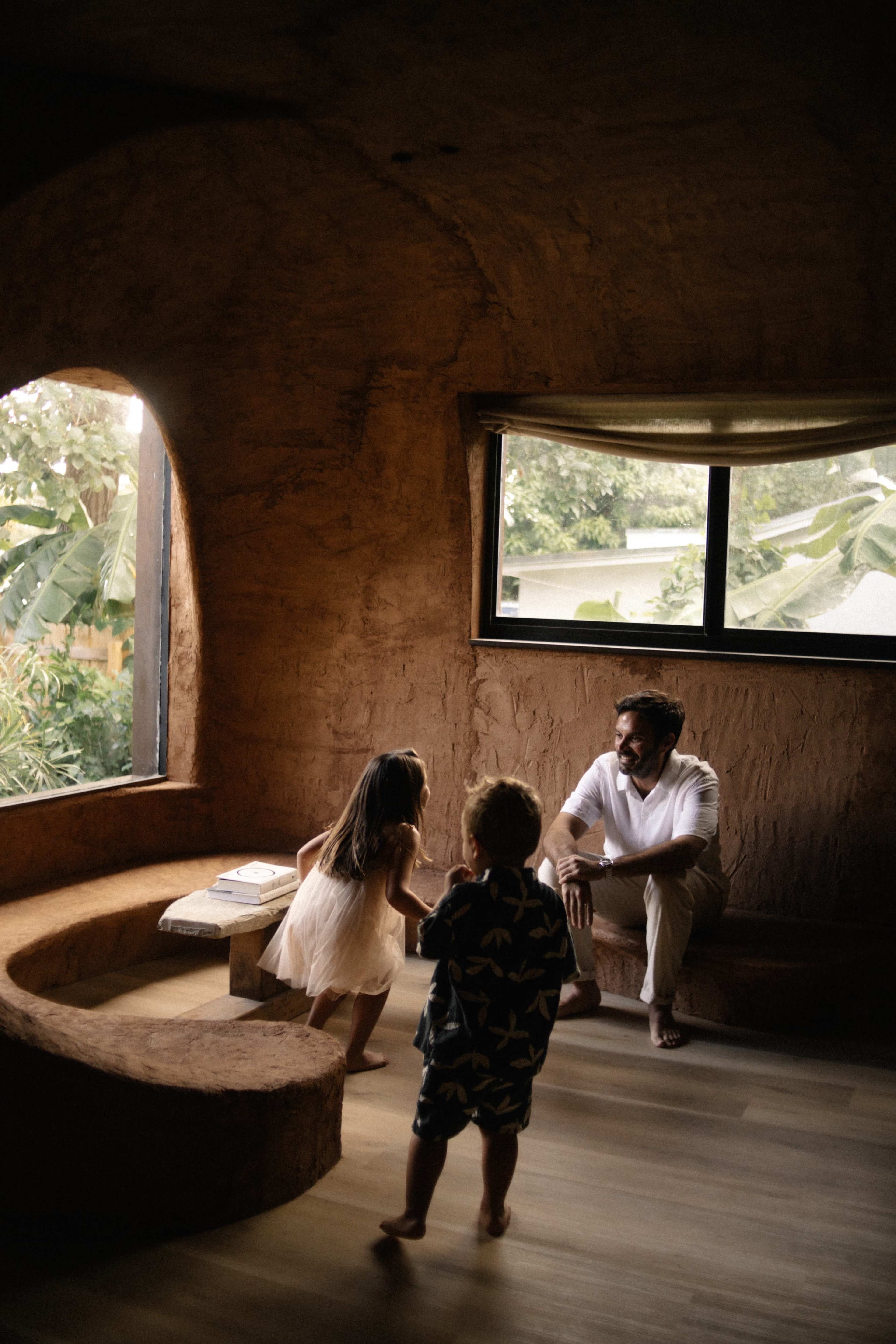 A father sits on the floor in a natural earthen room while two children play nearby.