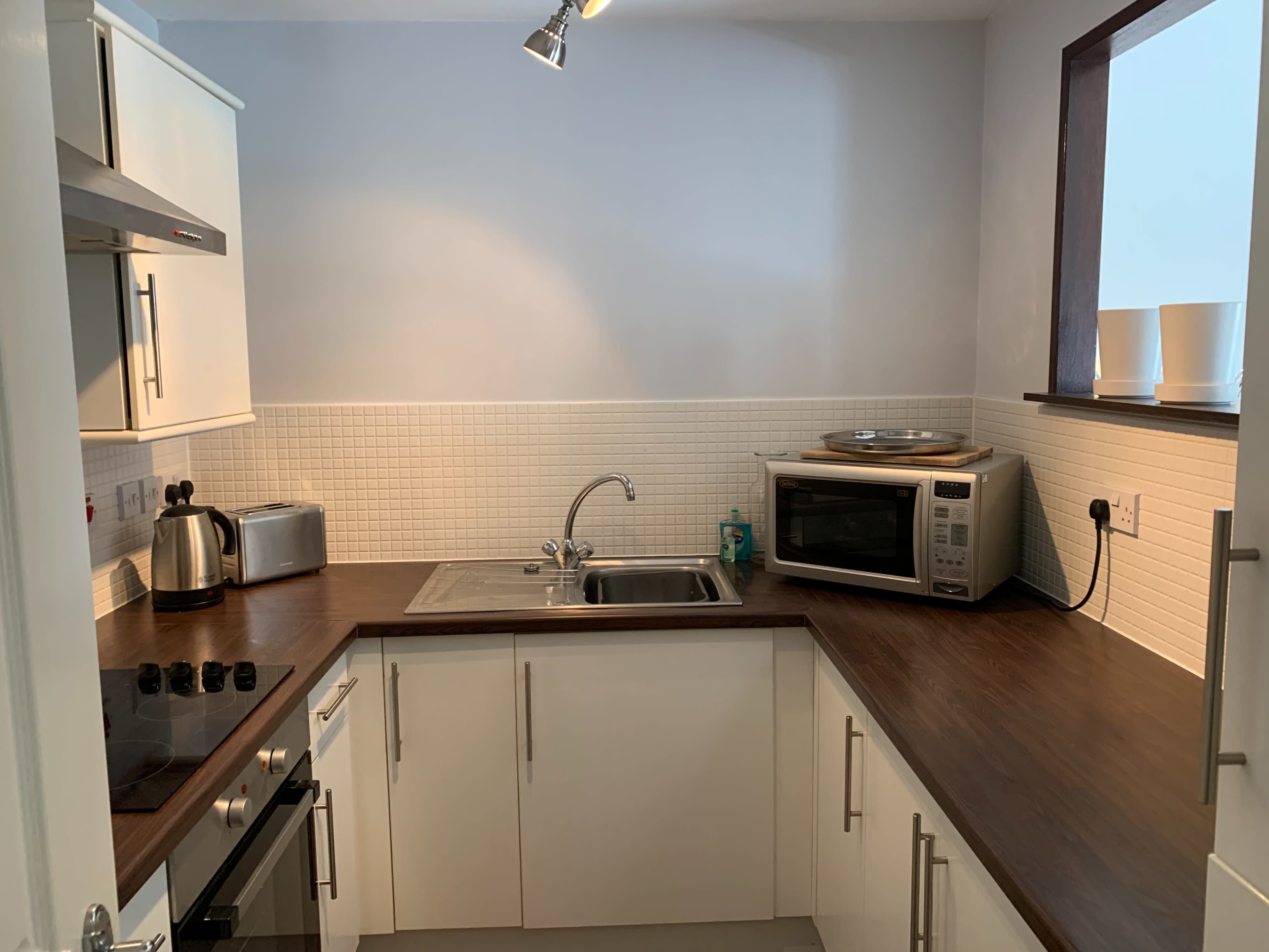 A kitchen with white cabinetry, a stainless steel sink, a microwave, and a countertop with a stove and kettle.