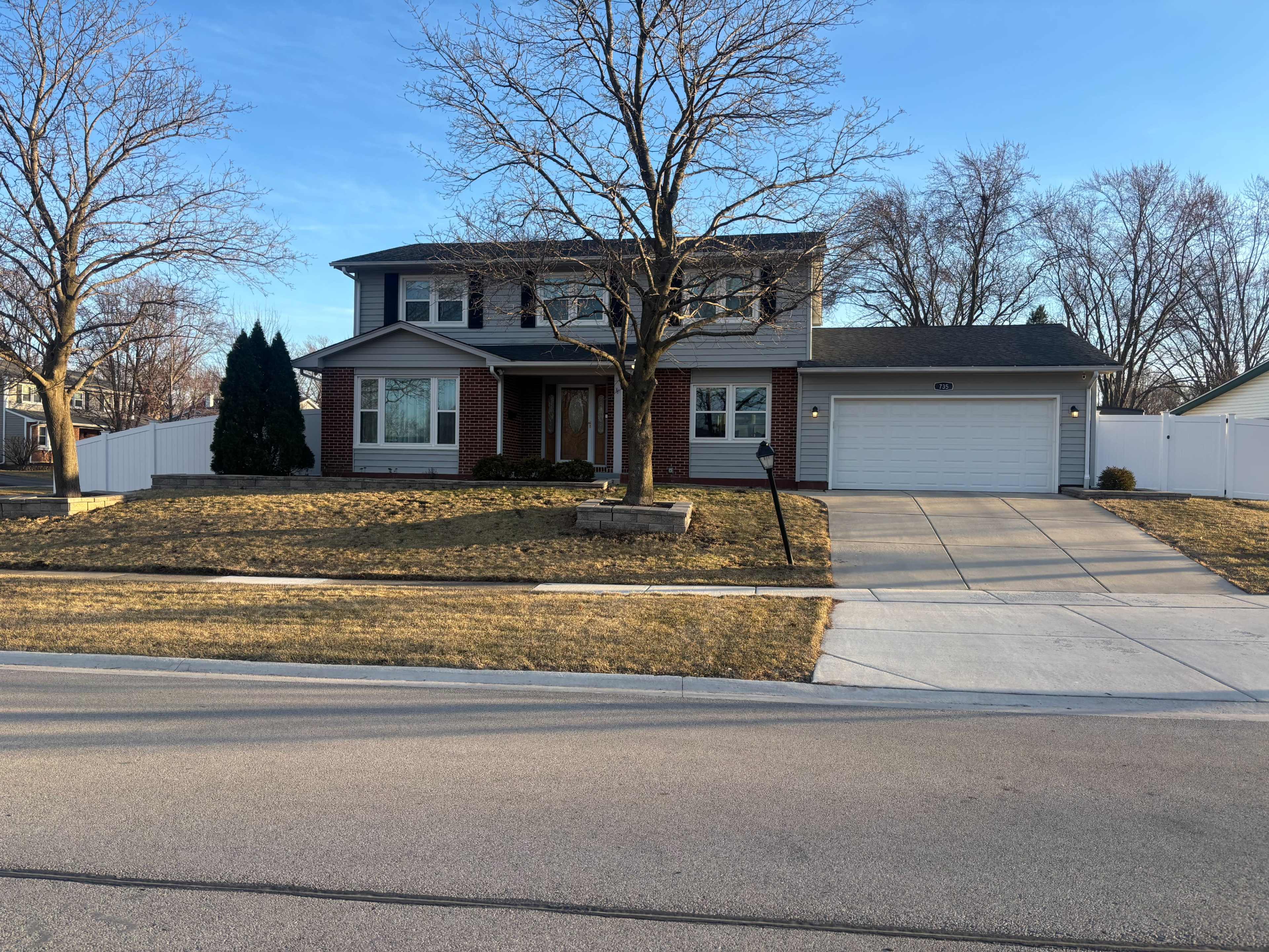 A two-story brick and siding house is located on a residential street with a manicured lawn and a driveway leading to a garage.