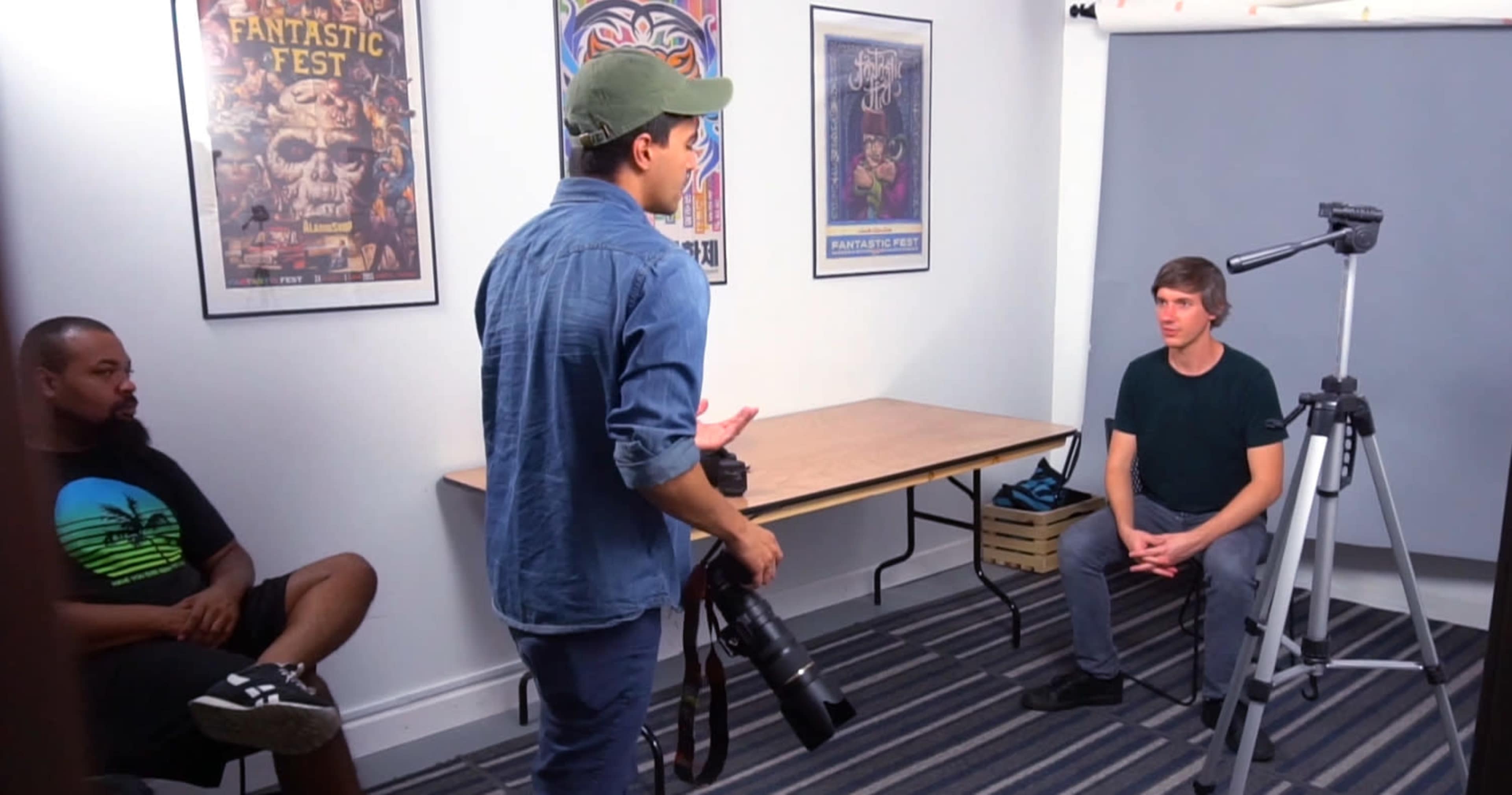 A photographer stands with a camera while directing a seated model in a small studio with posters on the walls.