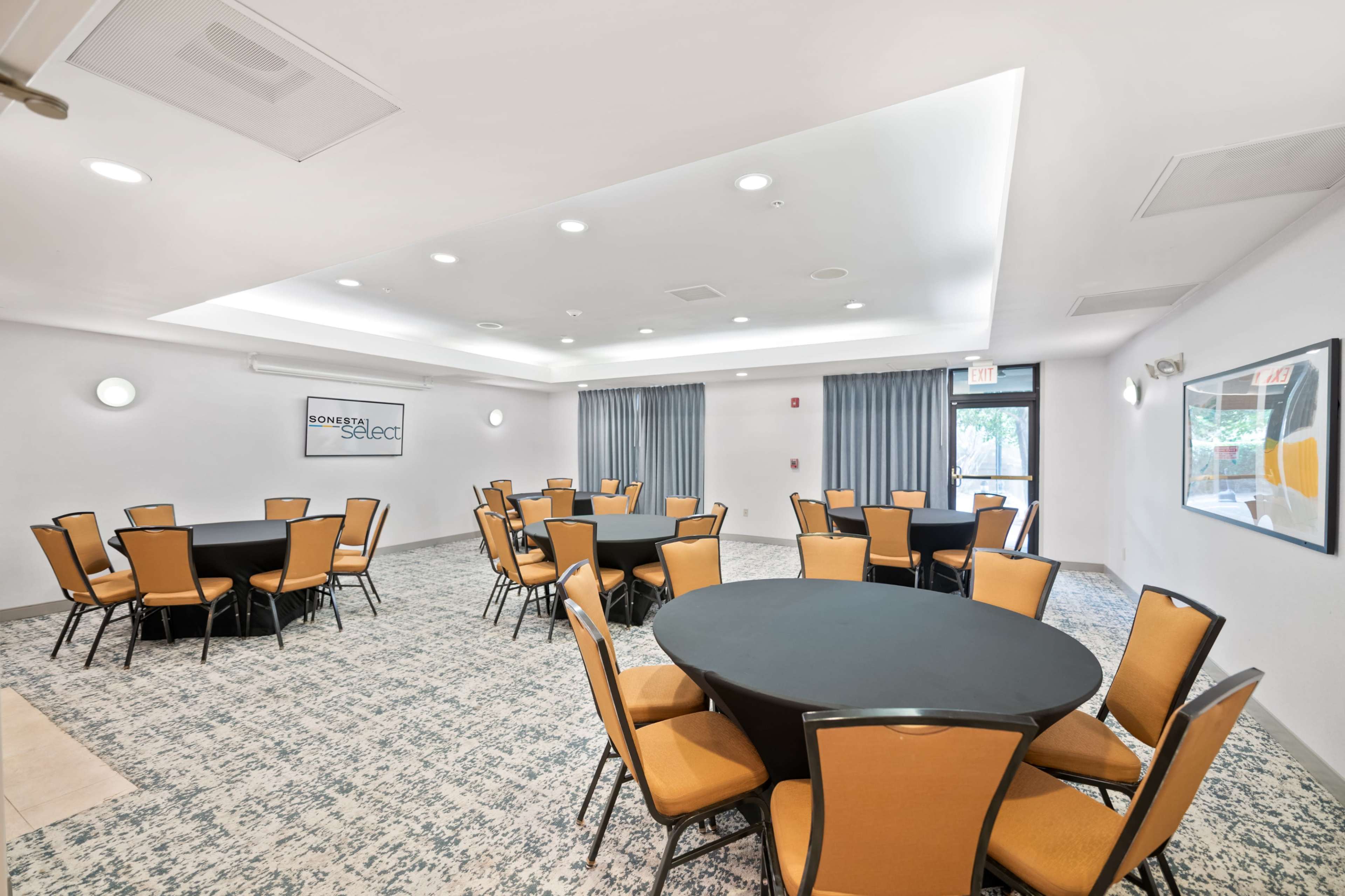 The image shows a well-lit meeting room featuring several round tables with orange and black chairs arranged neatly on a patterned carpet.