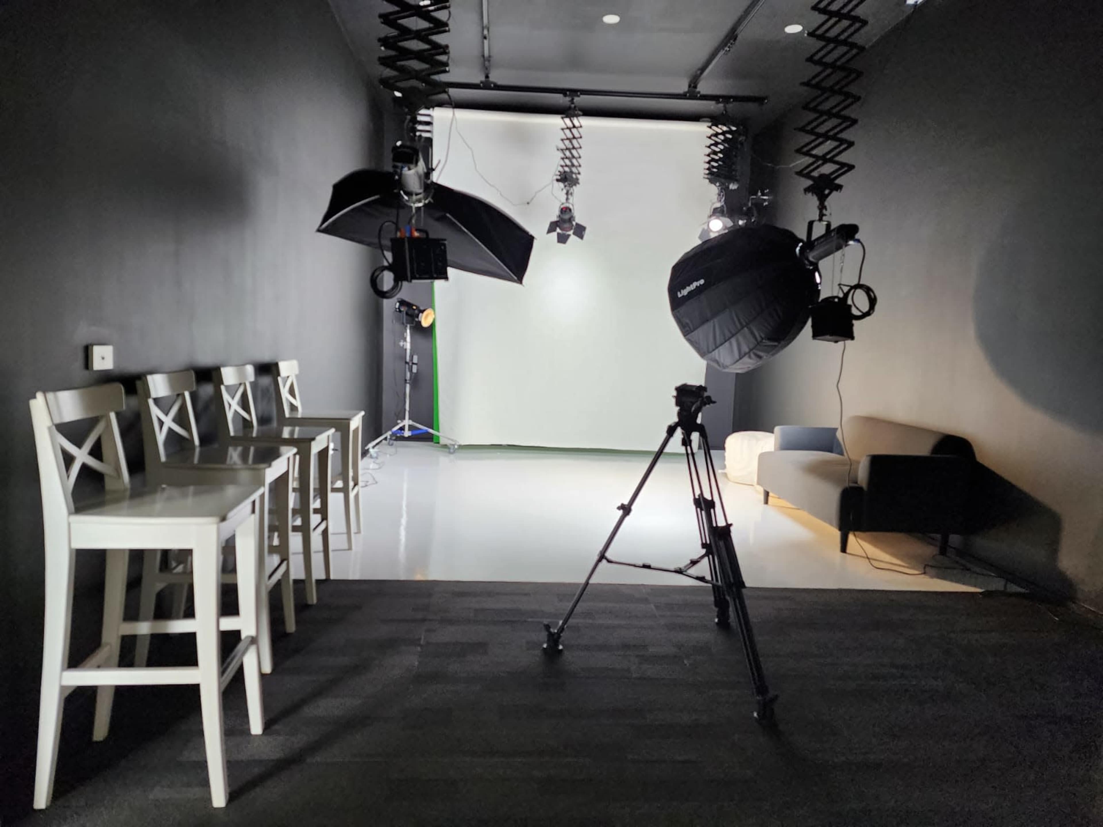 A studio space featuring a row of white barstools, a camera tripod, and lighting equipment facing a backdrop.