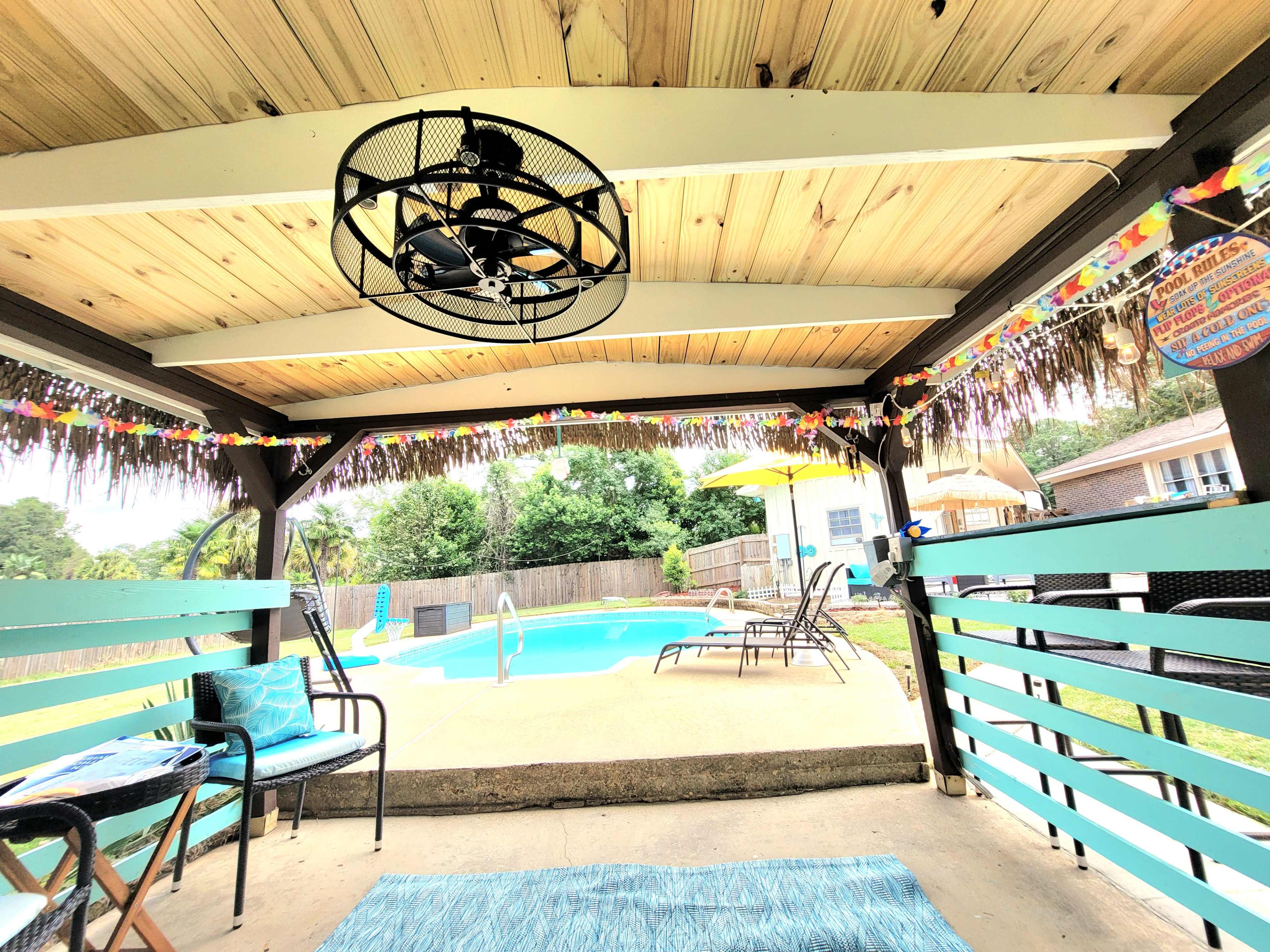 The image shows a covered outdoor patio with a ceiling fan, decorated with colorful streamers, overlooking a pool area with lounge chairs and umbrellas.