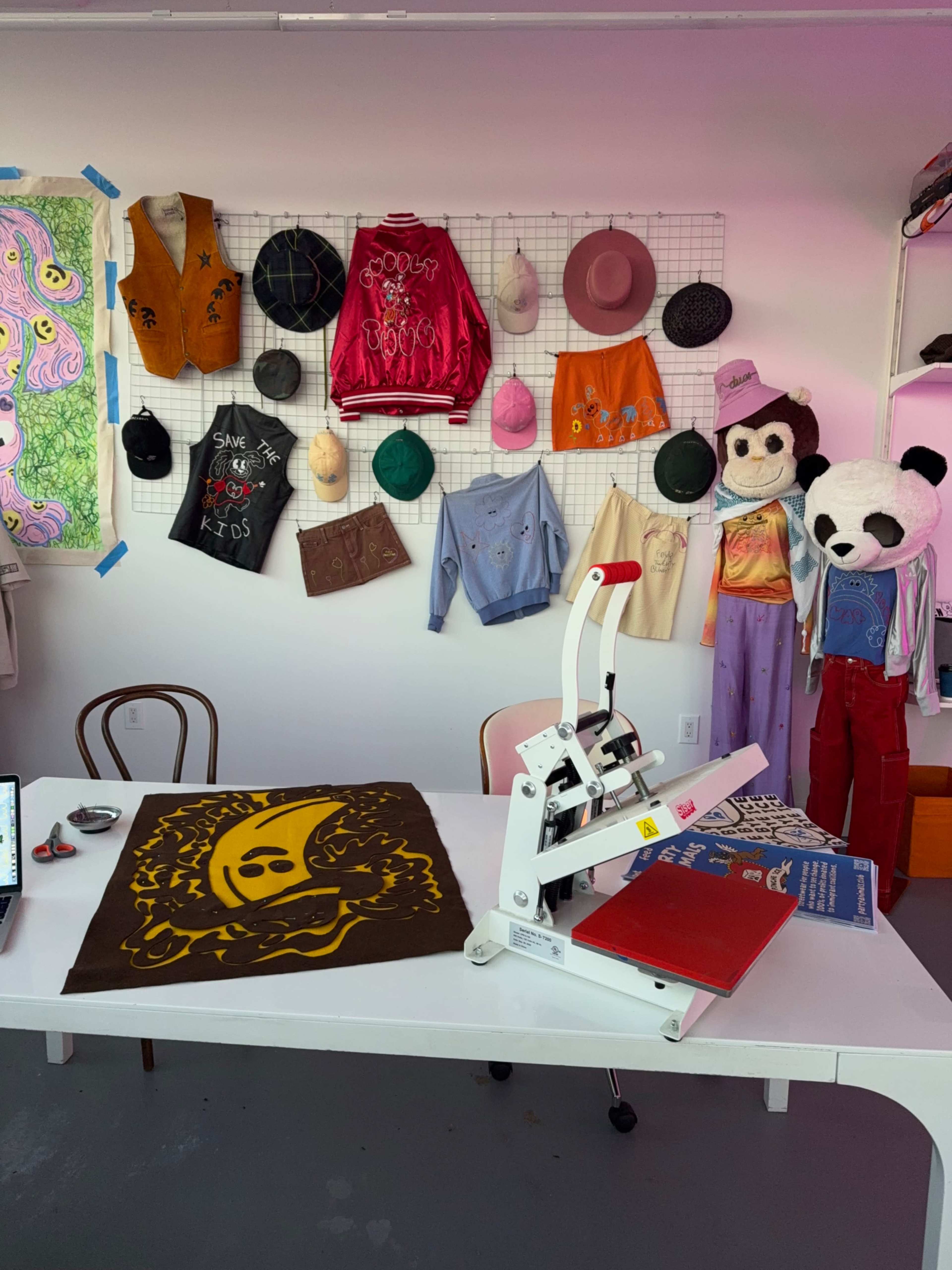 The image shows a workspace featuring a heat press machine on a table, surrounded by colorful clothing items and accessories hanging on a wall.