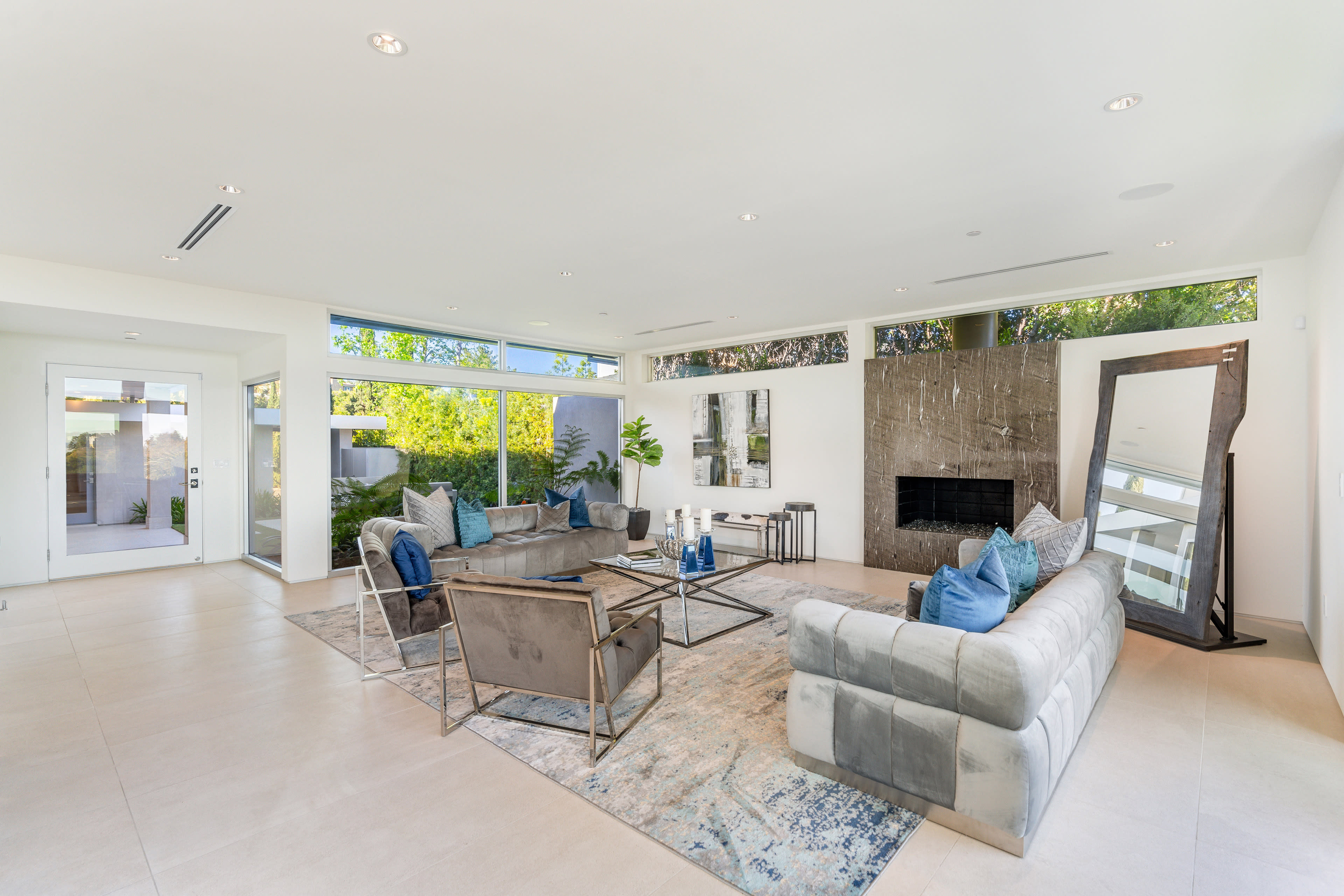 The image shows a modern living room featuring a sectional sofa, a coffee table, and large windows with a view of greenery.