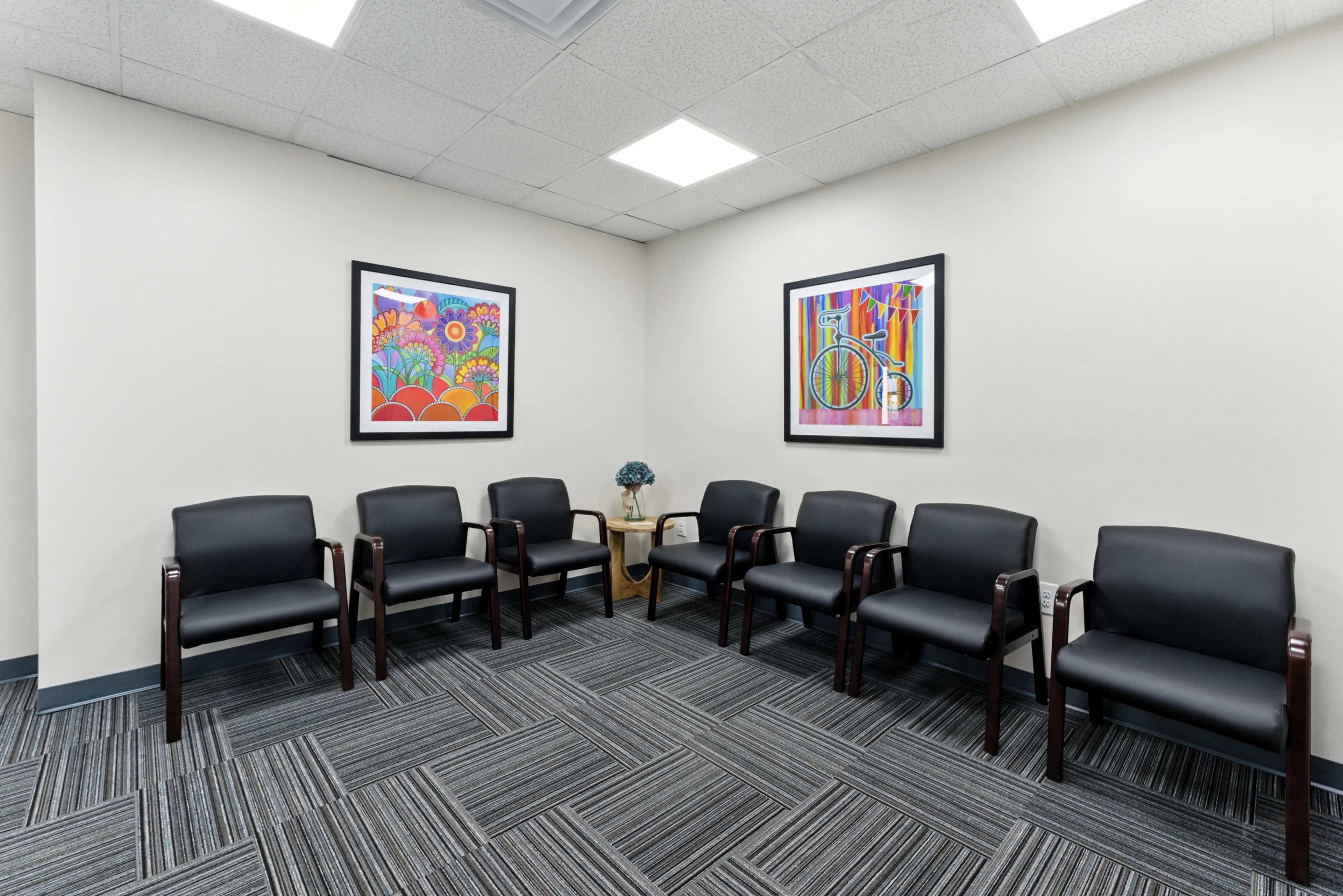 The image depicts a waiting area featuring six black chairs arranged in a semi-circle around a small table, with colorful artwork hanging on the walls.