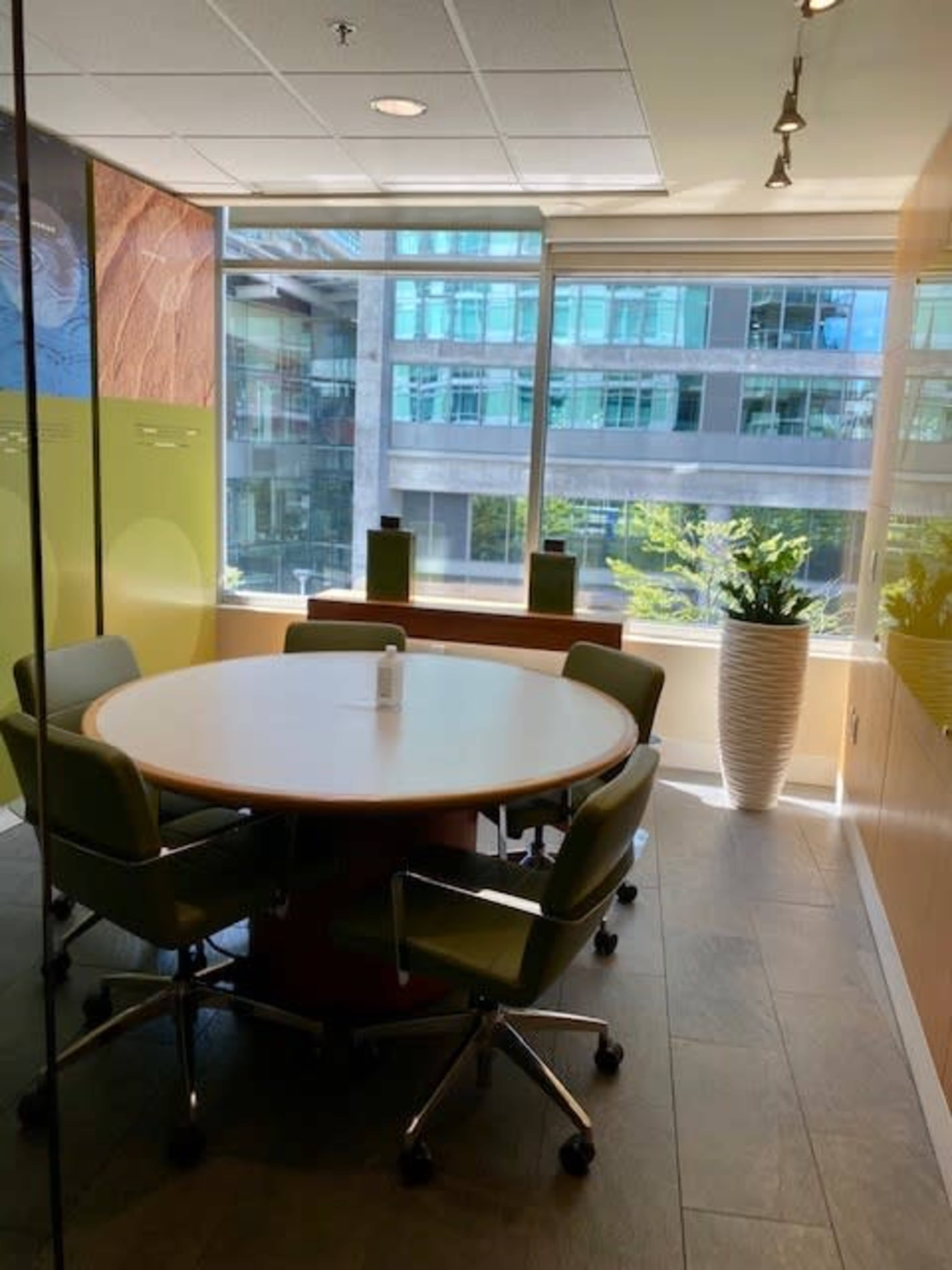 A modern conference room with a round table surrounded by green chairs, large windows providing natural light, and a decorative plant in a tall vase.