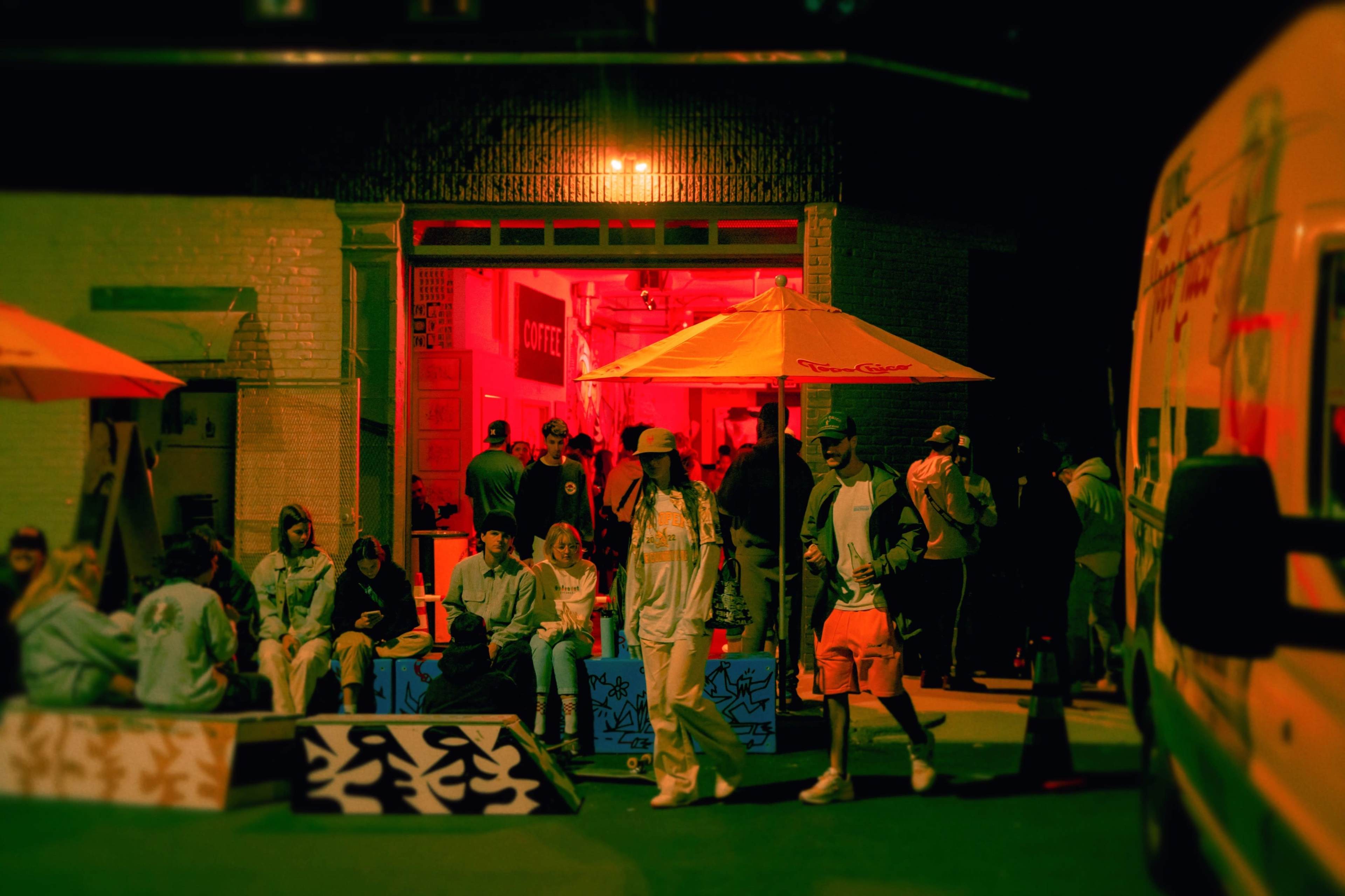 A group of people socializes outside a brightly lit coffee shop at night, with colorful umbrellas providing shade over seated patrons.