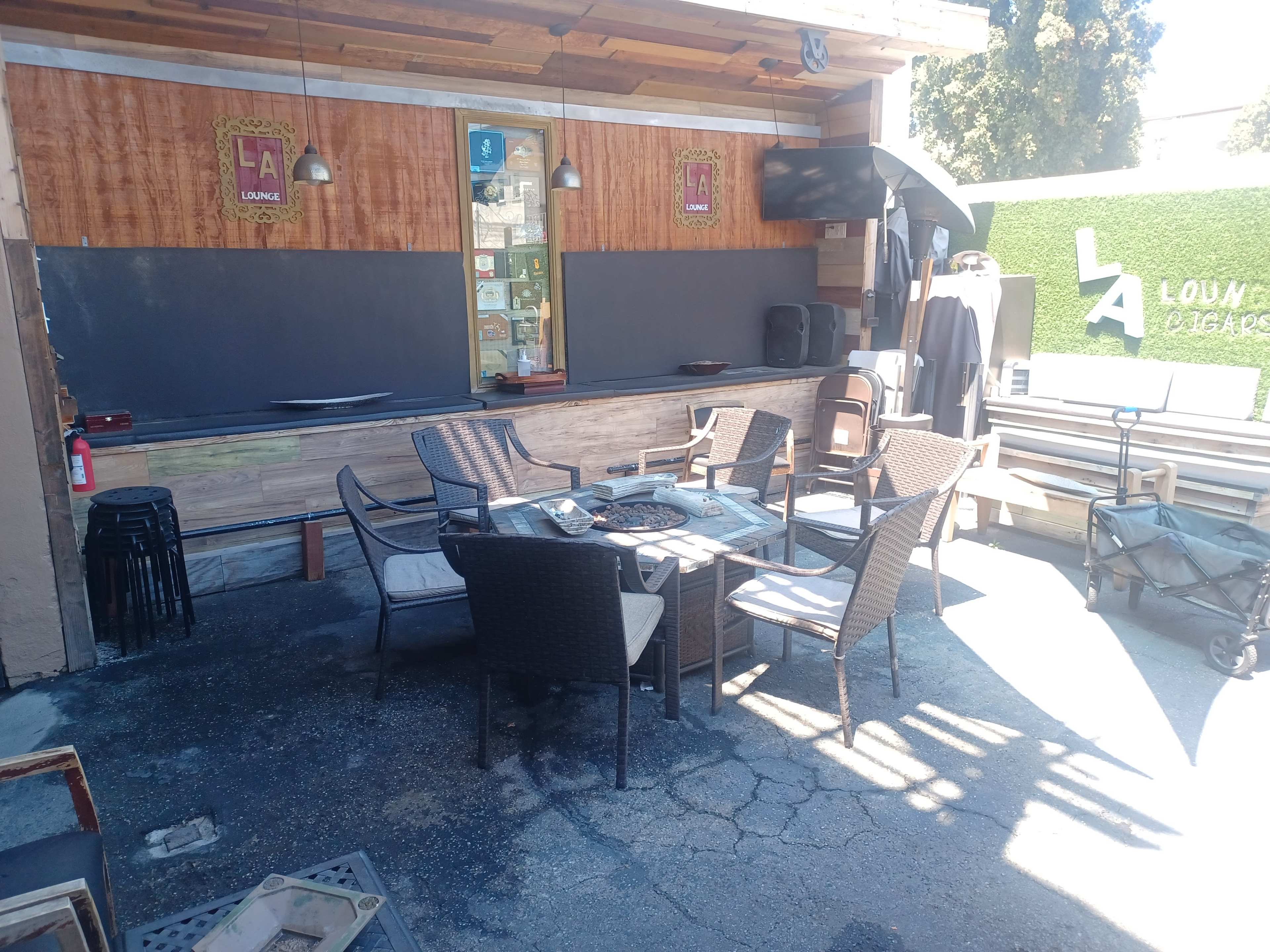 An outdoor lounge area with a table and several chairs arranged around it, adjacent to a bar counter and a backdrop featuring green plants.