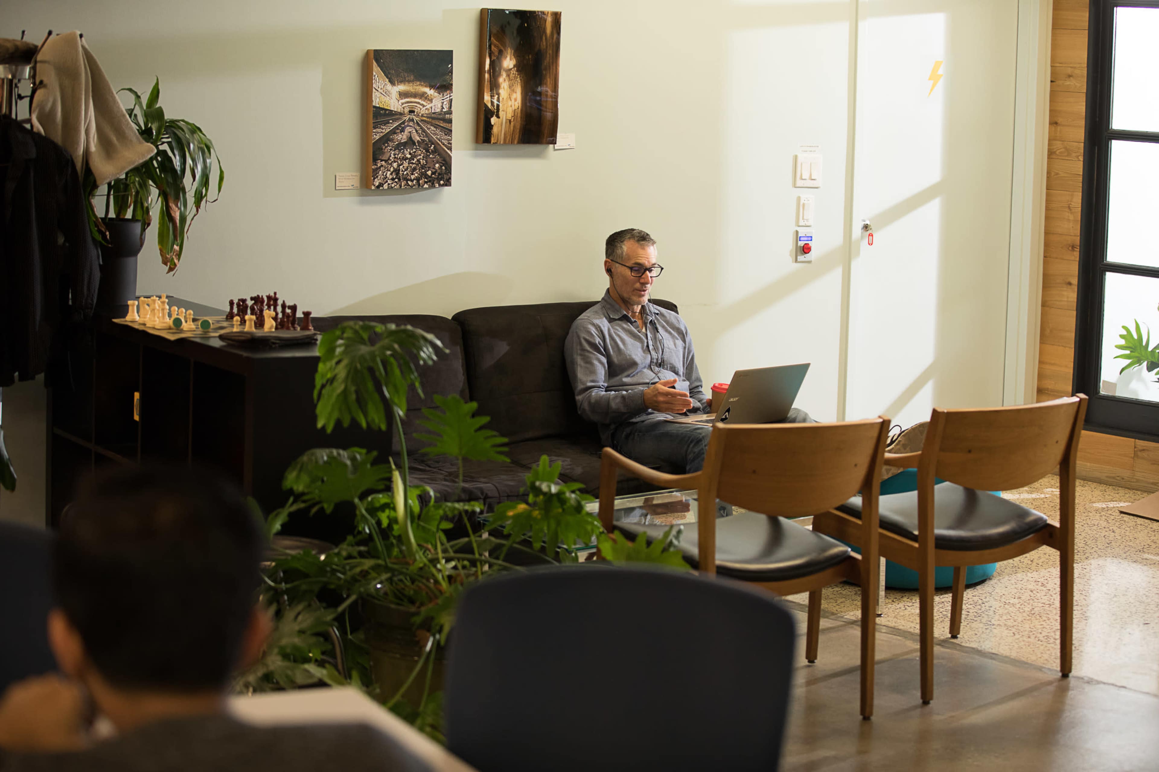 A man sits on a sofa working on a laptop in a room with plants and various seating arrangements.