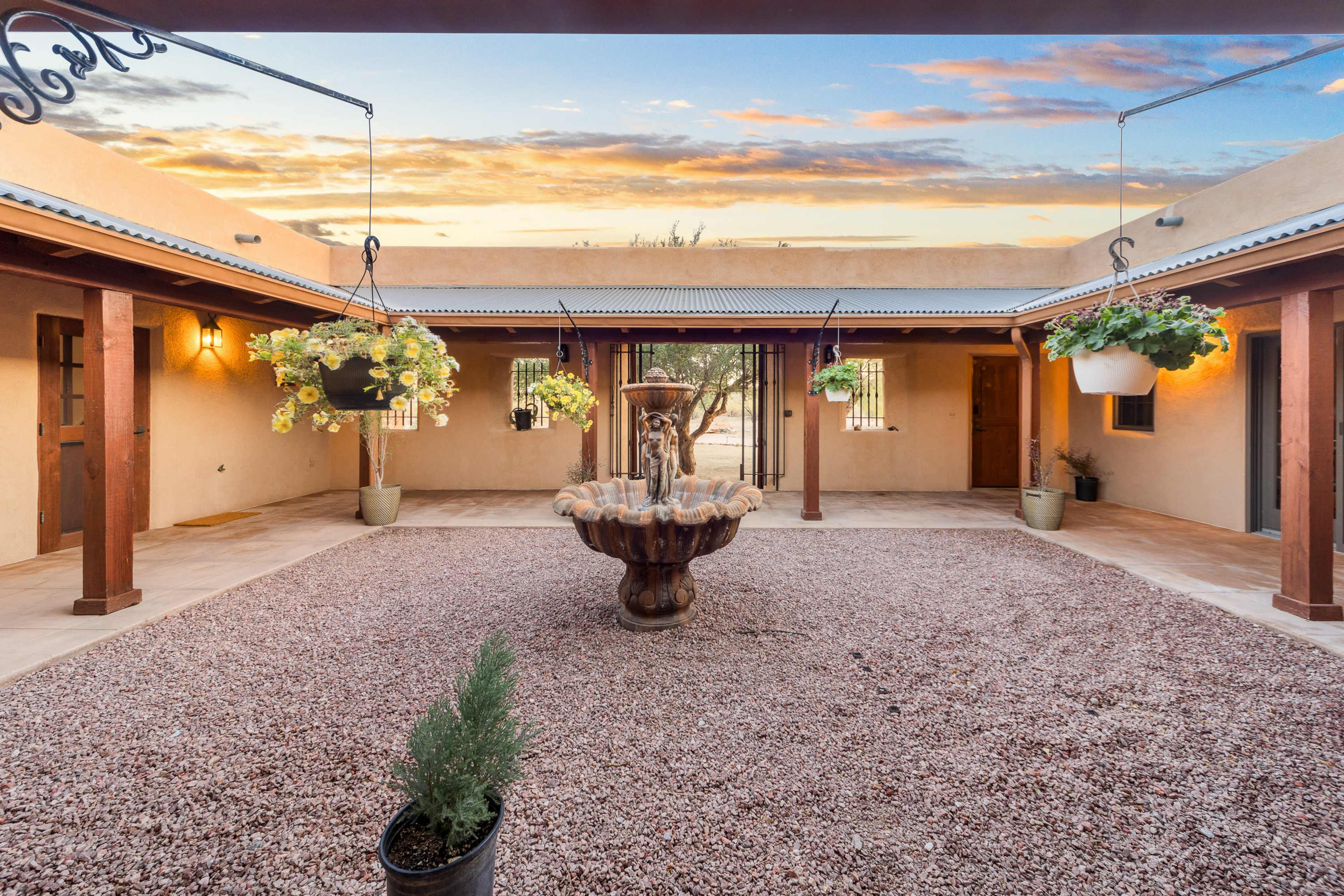 The image depicts a courtyard with a central fountain surrounded by potted plants and a gravel surface, framed by adobe-style buildings under a colorful sunset sky.