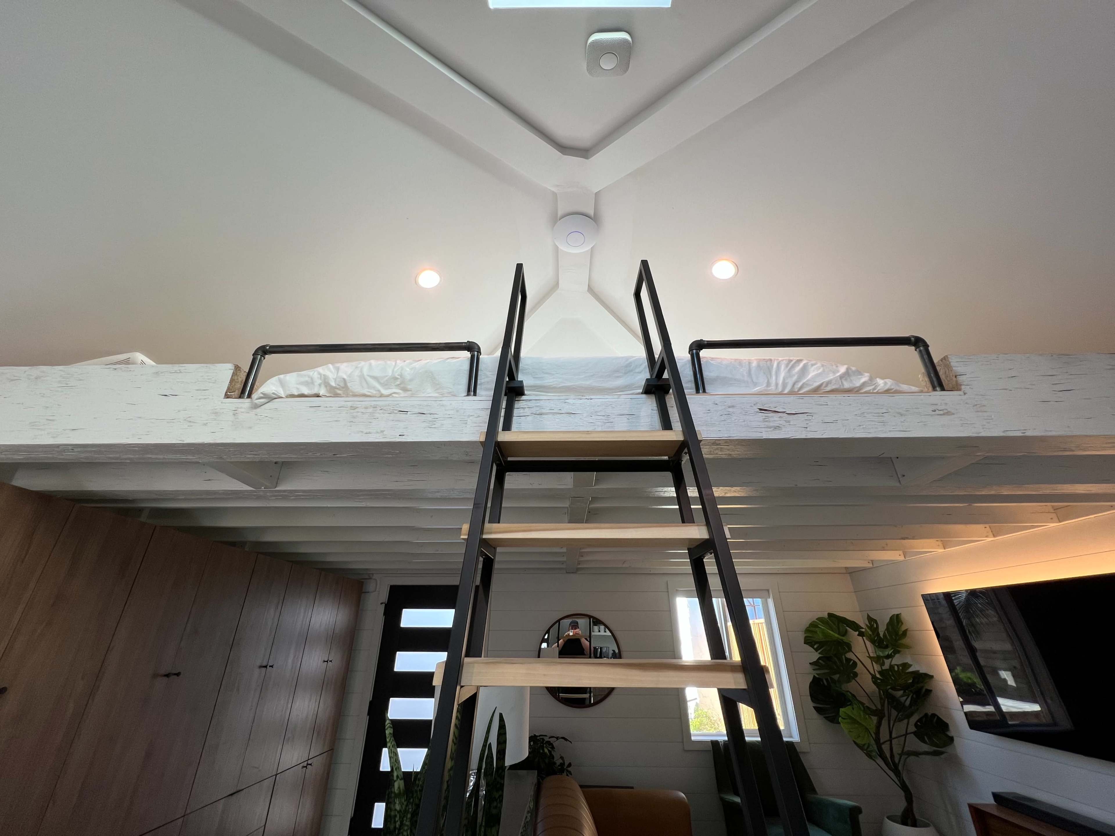 A wooden ladder leads to a lofted bed in a bright, modern room with large windows and a seating area below.