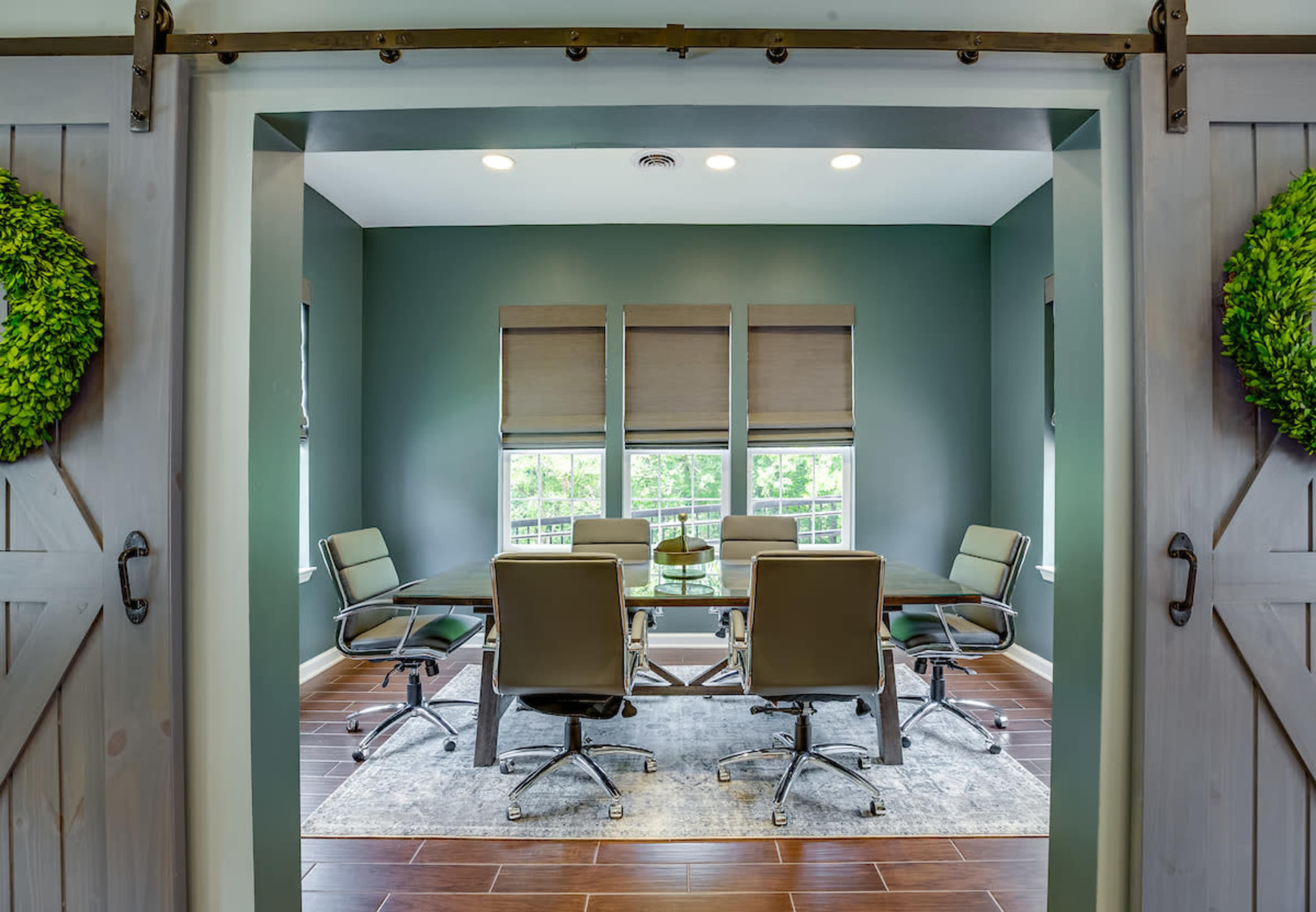 A conference room with a round glass table surrounded by six leather office chairs and large windows, framed by barn-style doors.