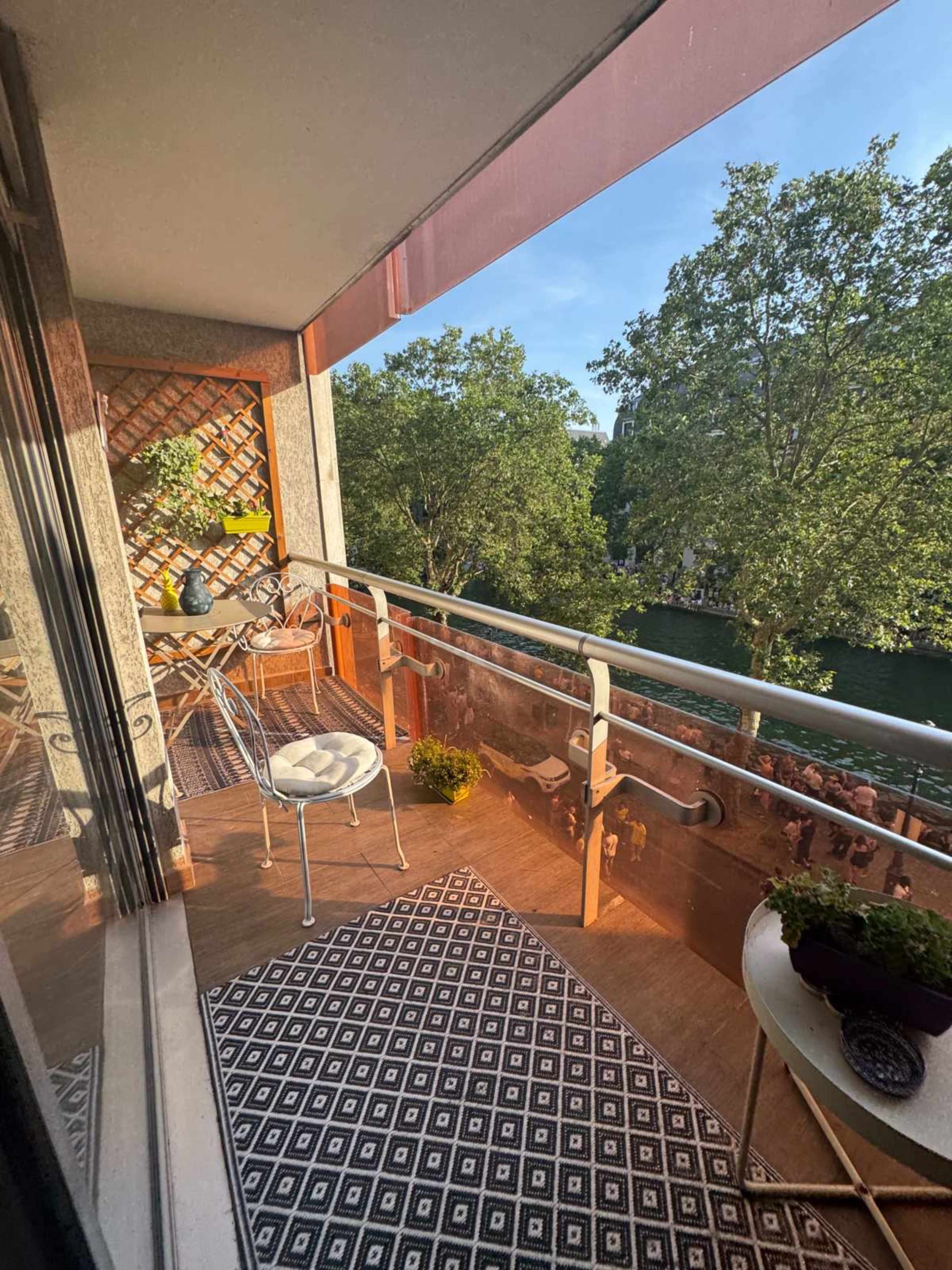 The image shows a balcony with a patterned rug, a small table and chair set, and potted plants, overlooking a tree-lined area.