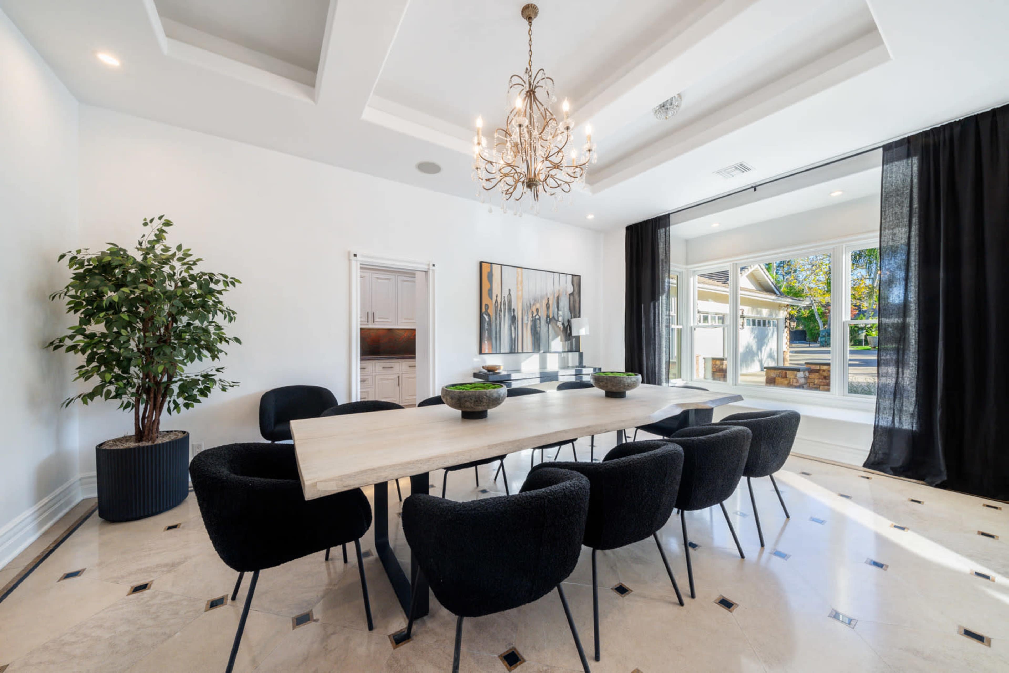 A large dining table surrounded by black upholstered chairs is set in a bright room with a chandelier and a window overlooking an outdoor space.