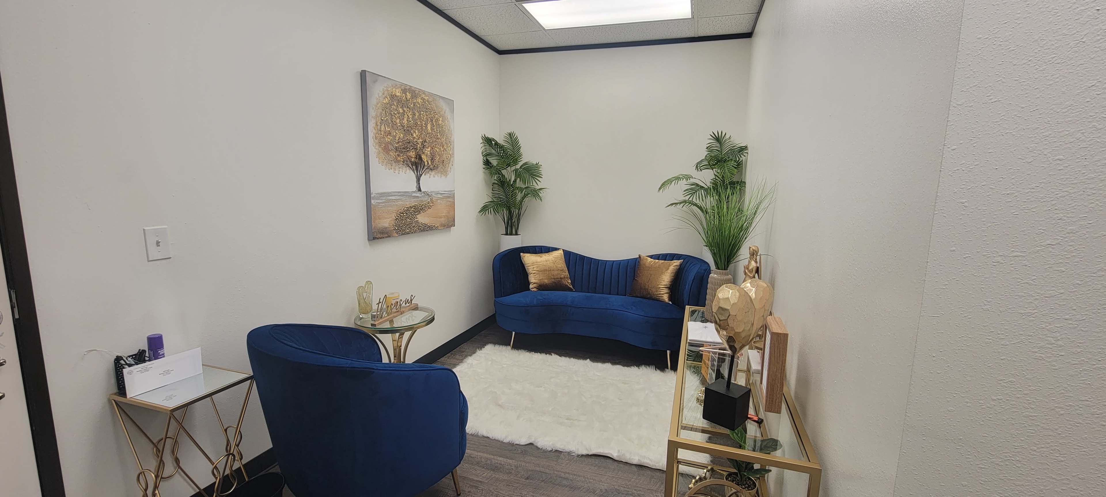 A small, elegantly arranged waiting area with a blue upholstered sofa, a matching chair, decorative plants, and a wall art featuring a tree.