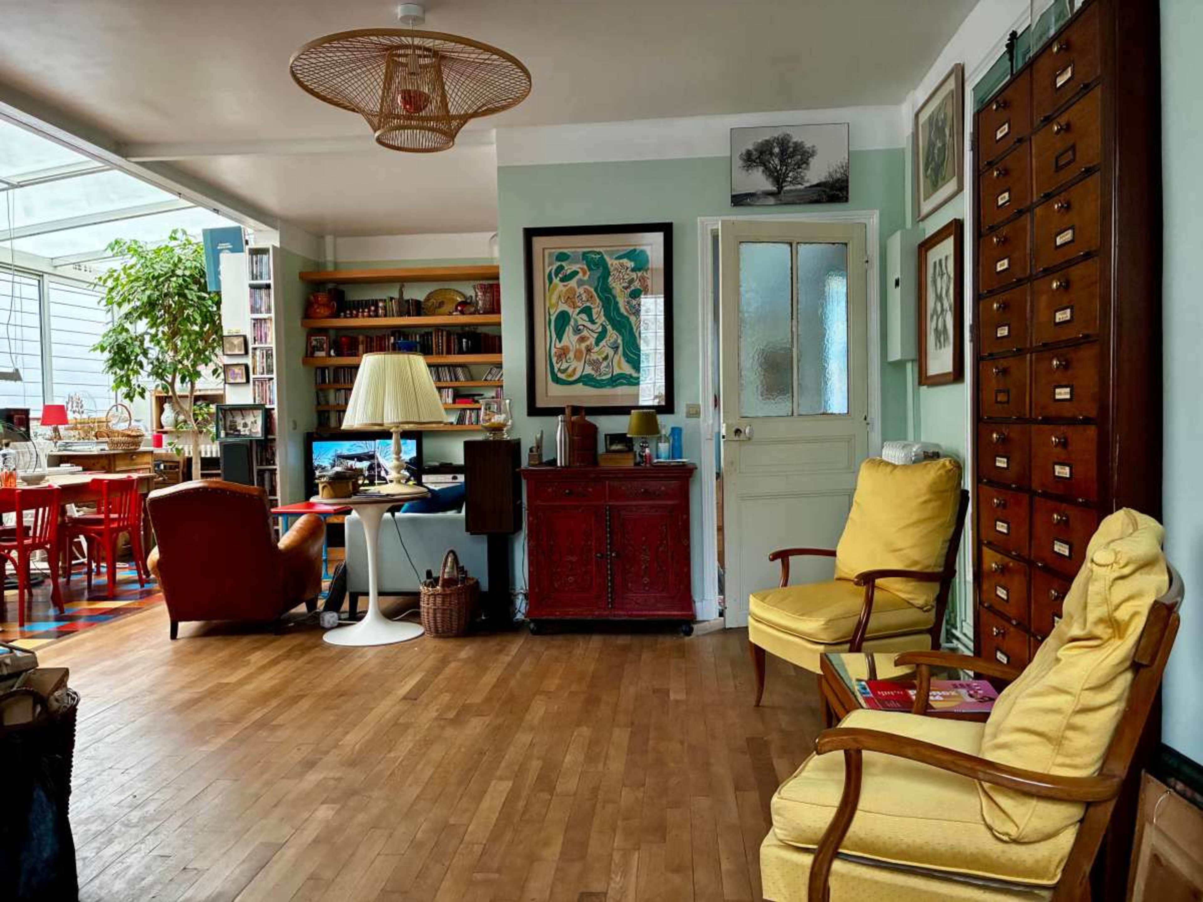 The image shows a cozy living room featuring two yellow armchairs, a wooden floor, a red cabinet, and bookshelves filled with books and decorative items.