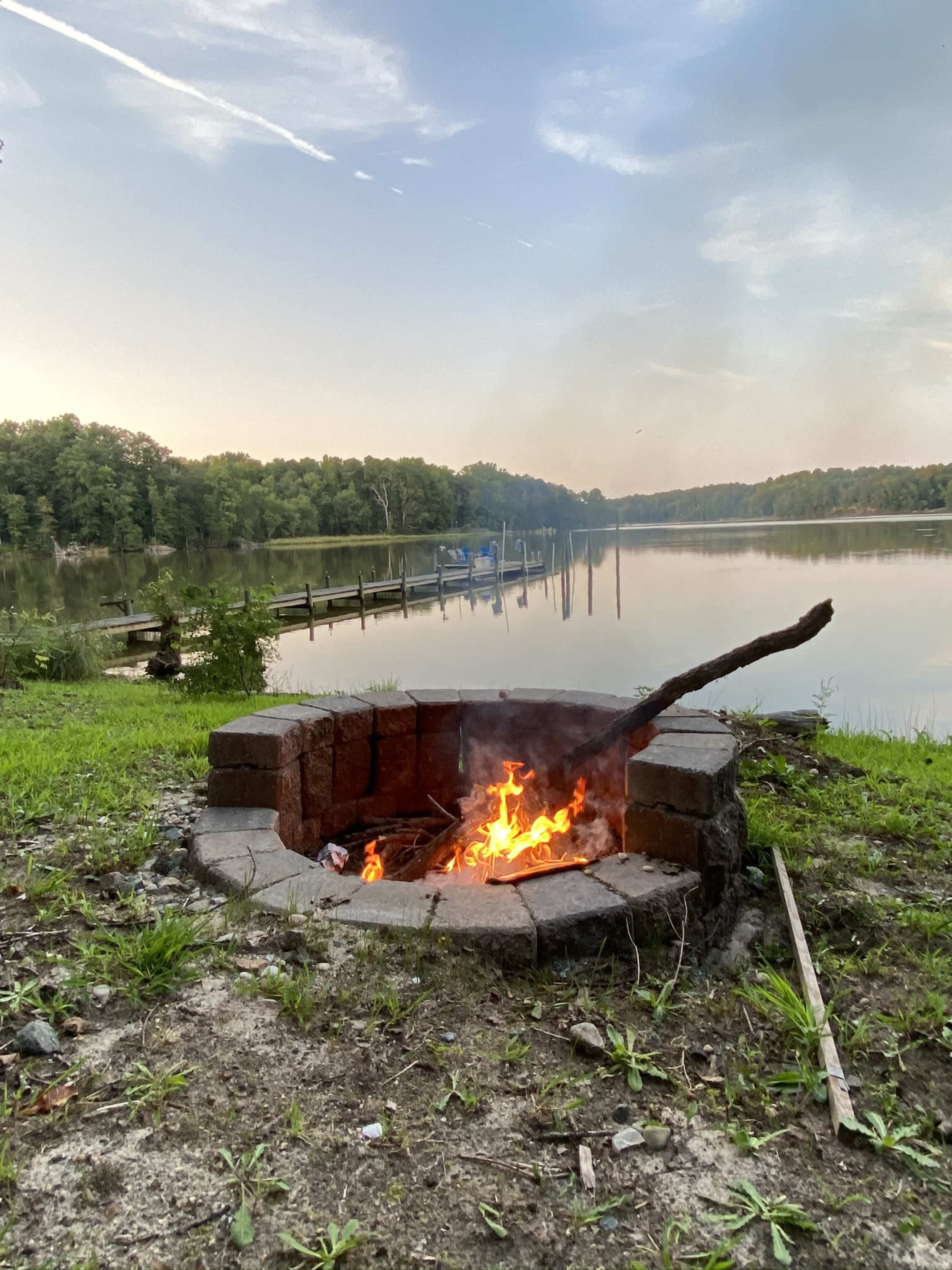 A stone fire pit with a flickering fire is situated near a calm lake, surrounded by green grass and trees, with a pier visible in the background.