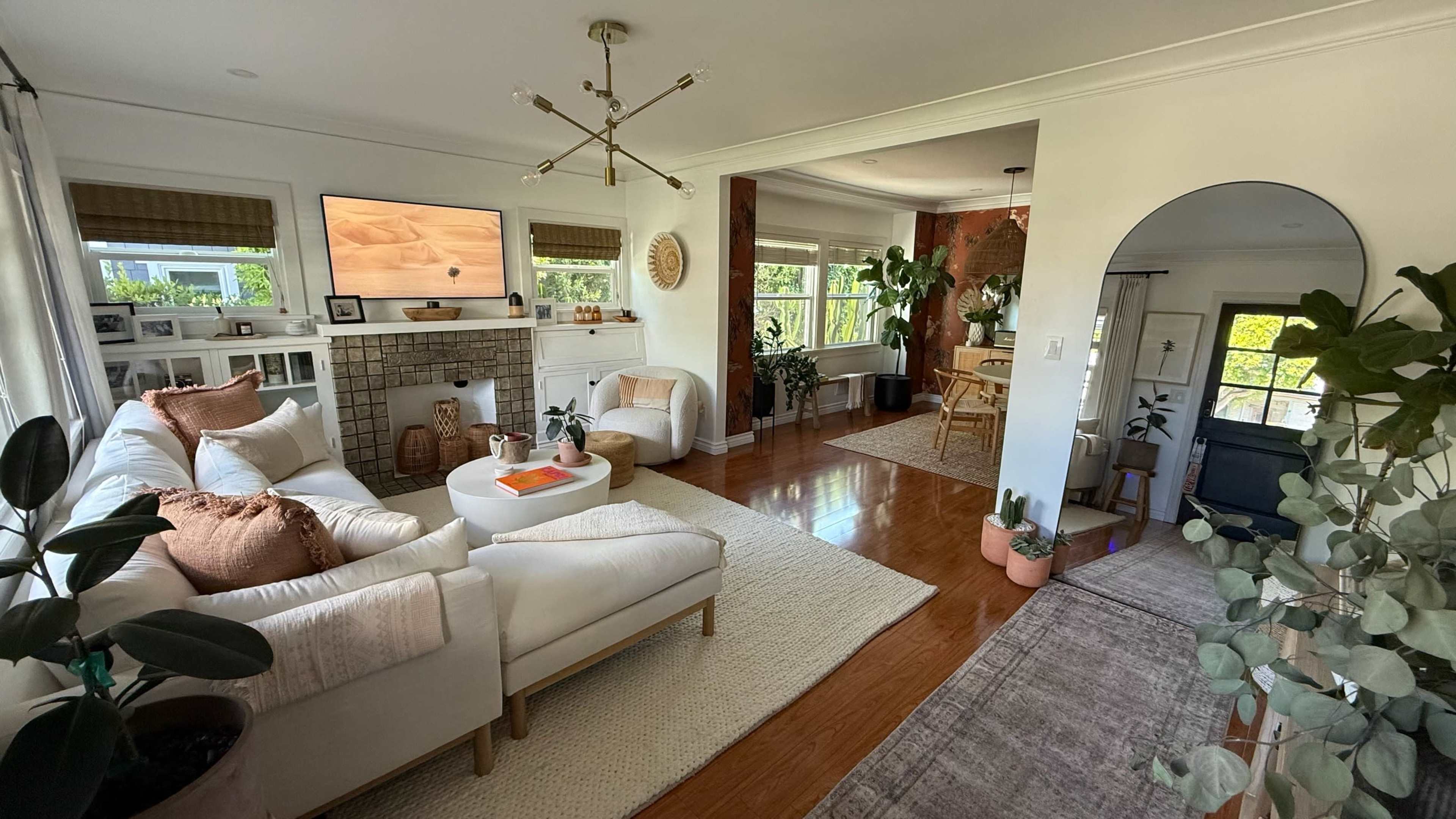 The image shows a brightly lit living room with a white sofa, armchair, coffee table, and plants, leading into a dining area.