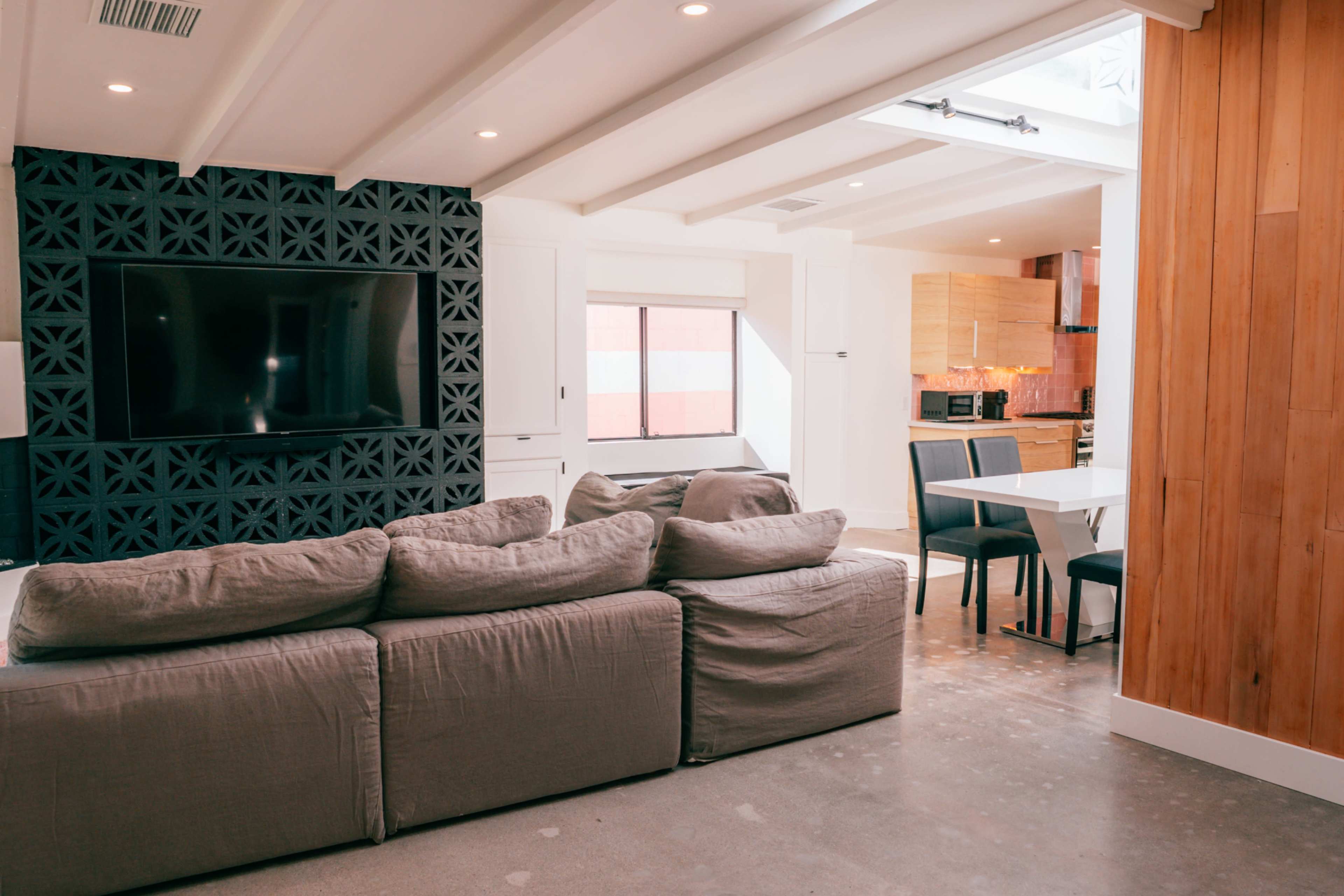 The image shows a modern living space featuring a large sectional sofa, a black patterned wall with a television, and an adjacent dining area with a white table and chairs.