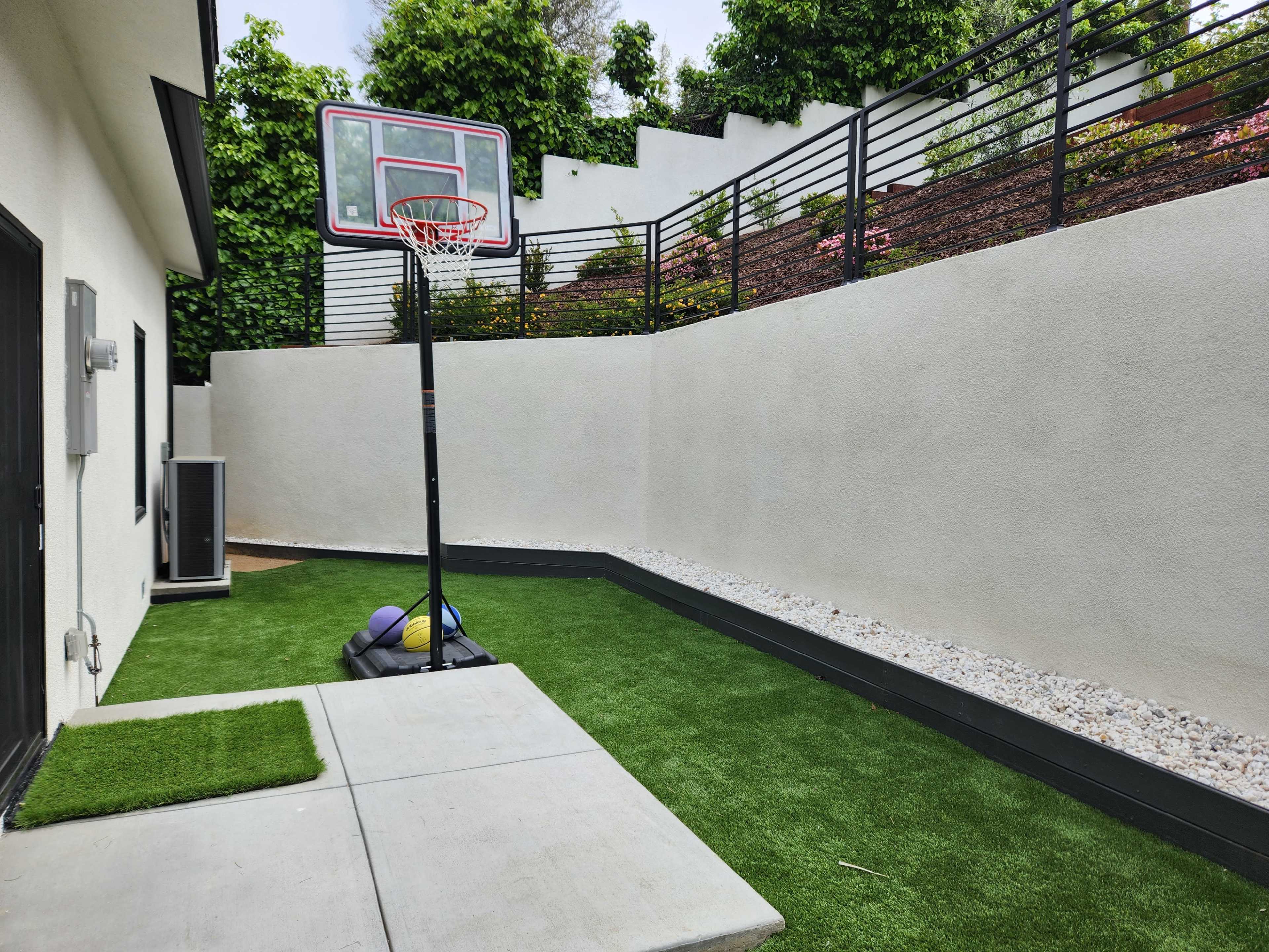 A gray concrete walkway leads to an empty basketball hoop beside a small patch of artificial grass and decorative rocks along a wall.