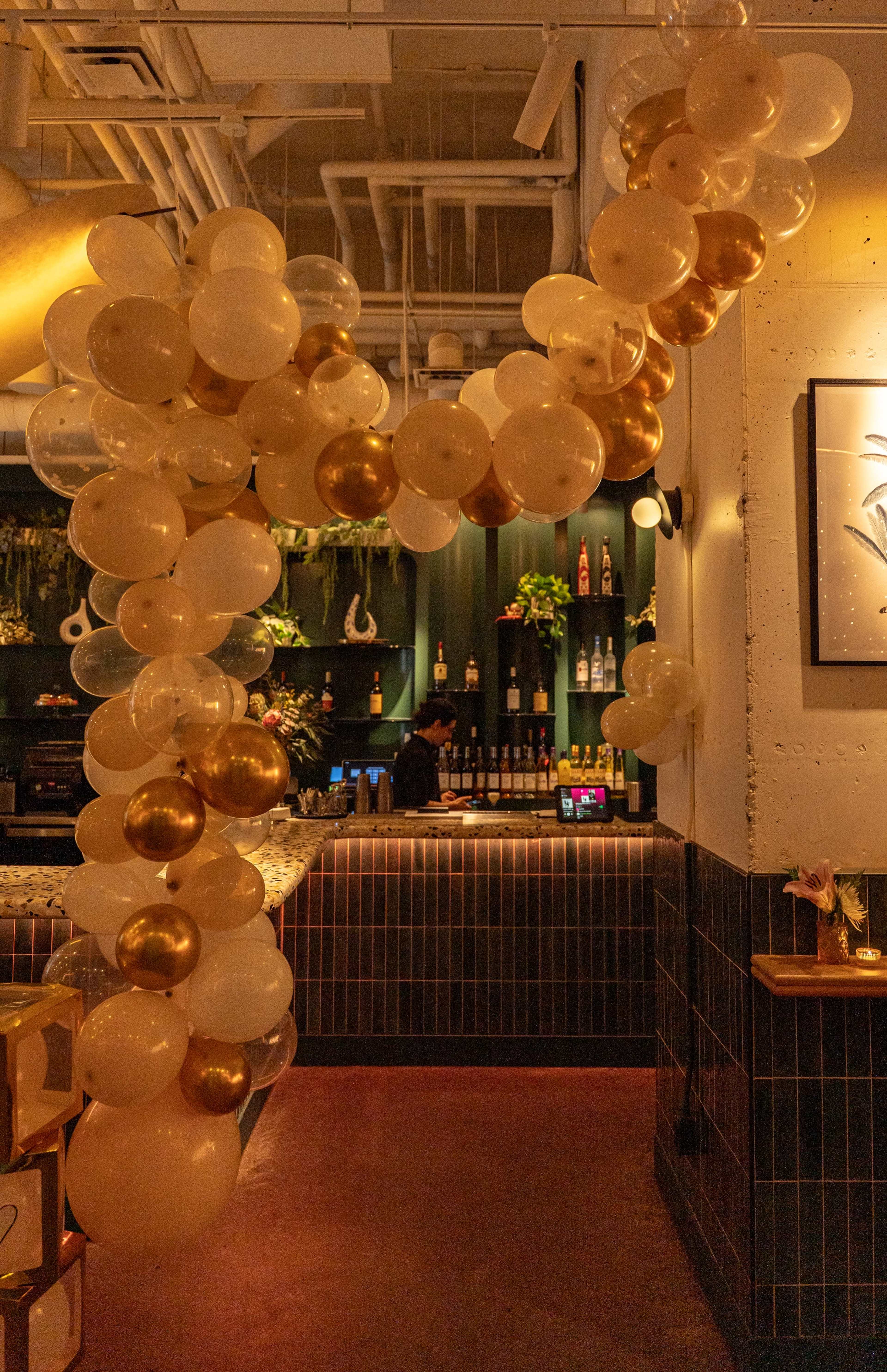 A large balloon arch made of white and gold balloons frames the entrance to a bar area with shelves of liquor in the background.