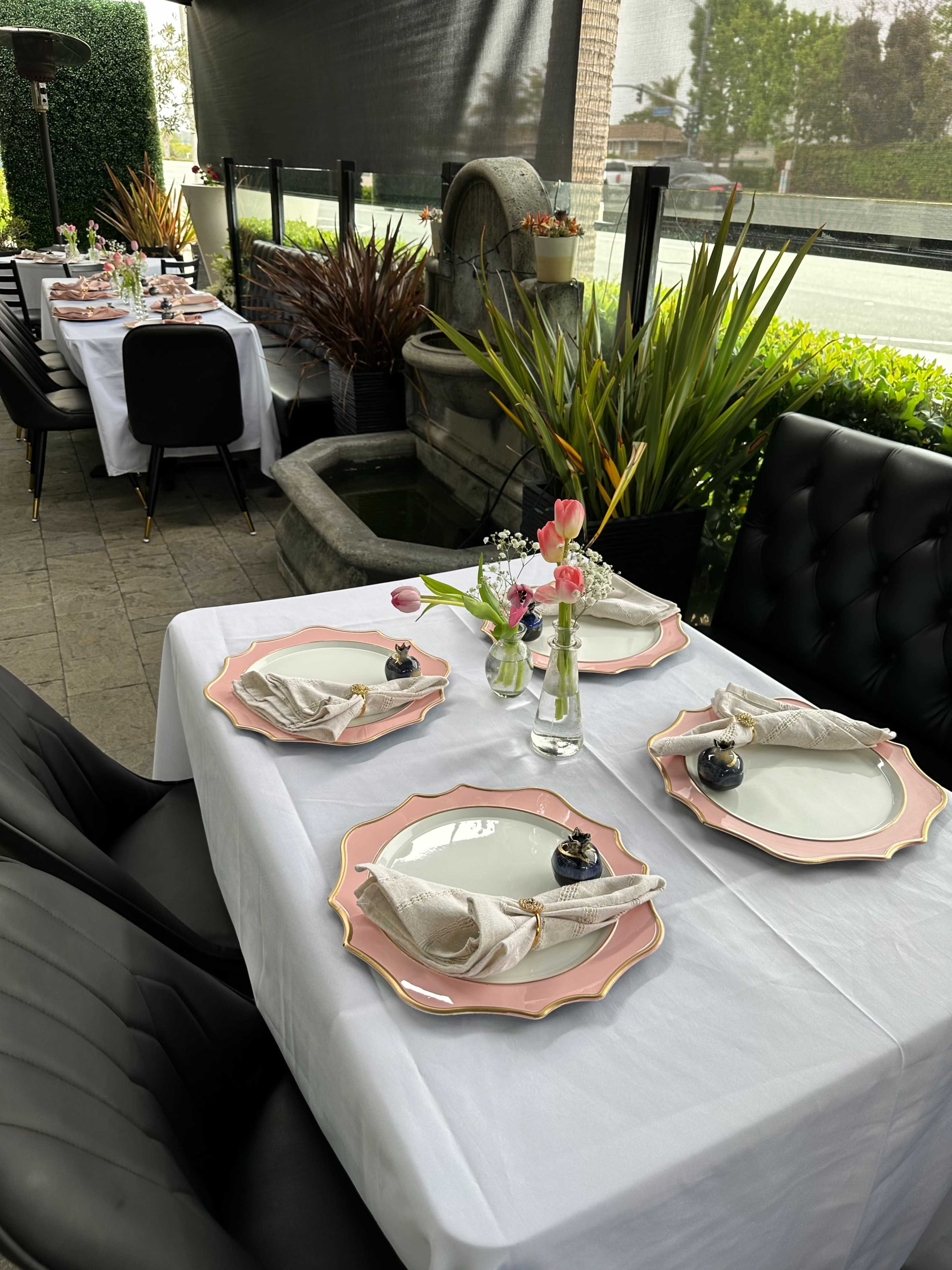 A dining table is set with pink rimmed plates, folded napkins, and small flower arrangements, surrounded by greenery and a decorative fountain in a cozy outdoor setting.