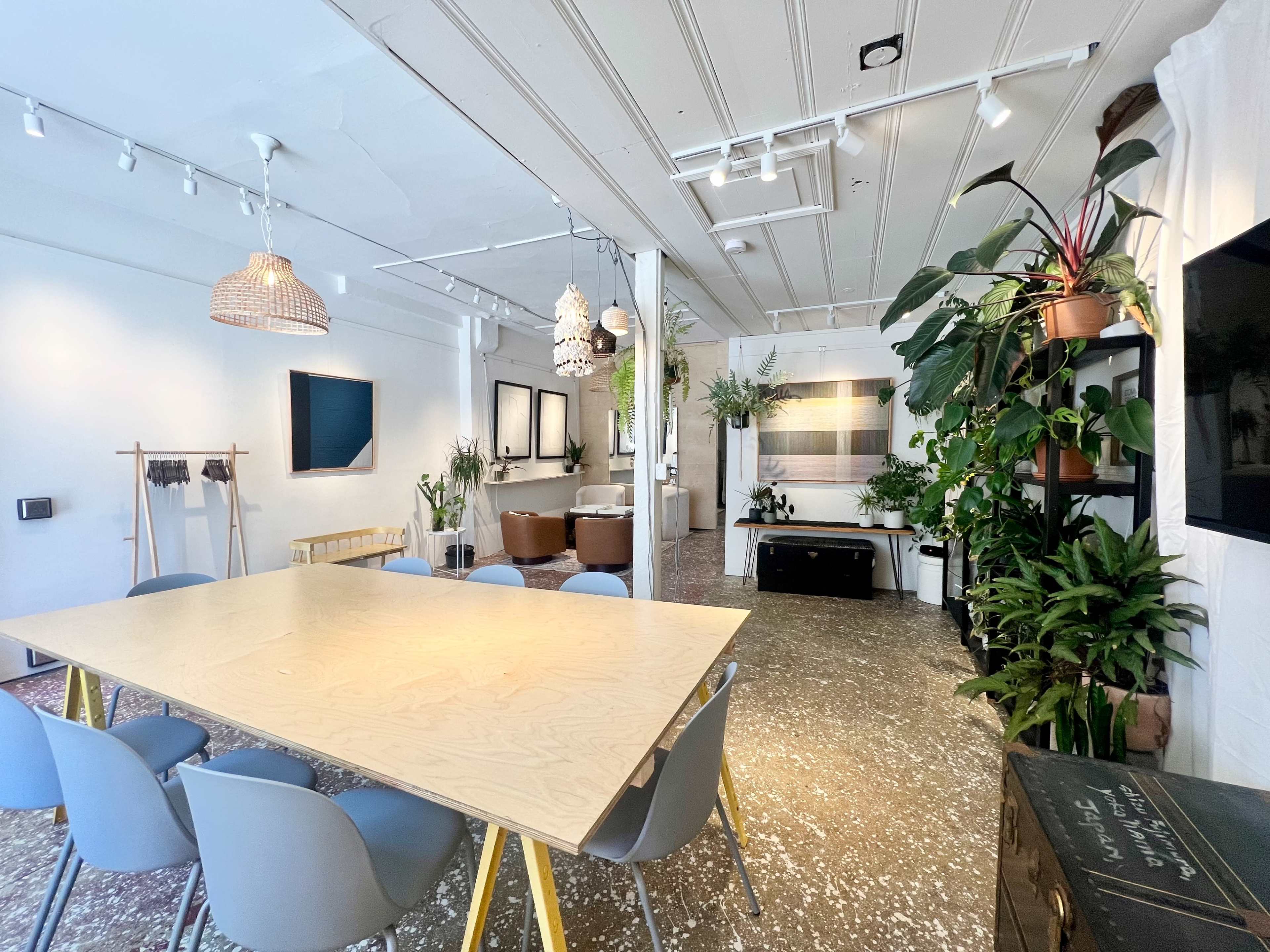 A spacious, modern interior features a large wooden table surrounded by blue chairs, with plants and artwork arranged throughout the room.