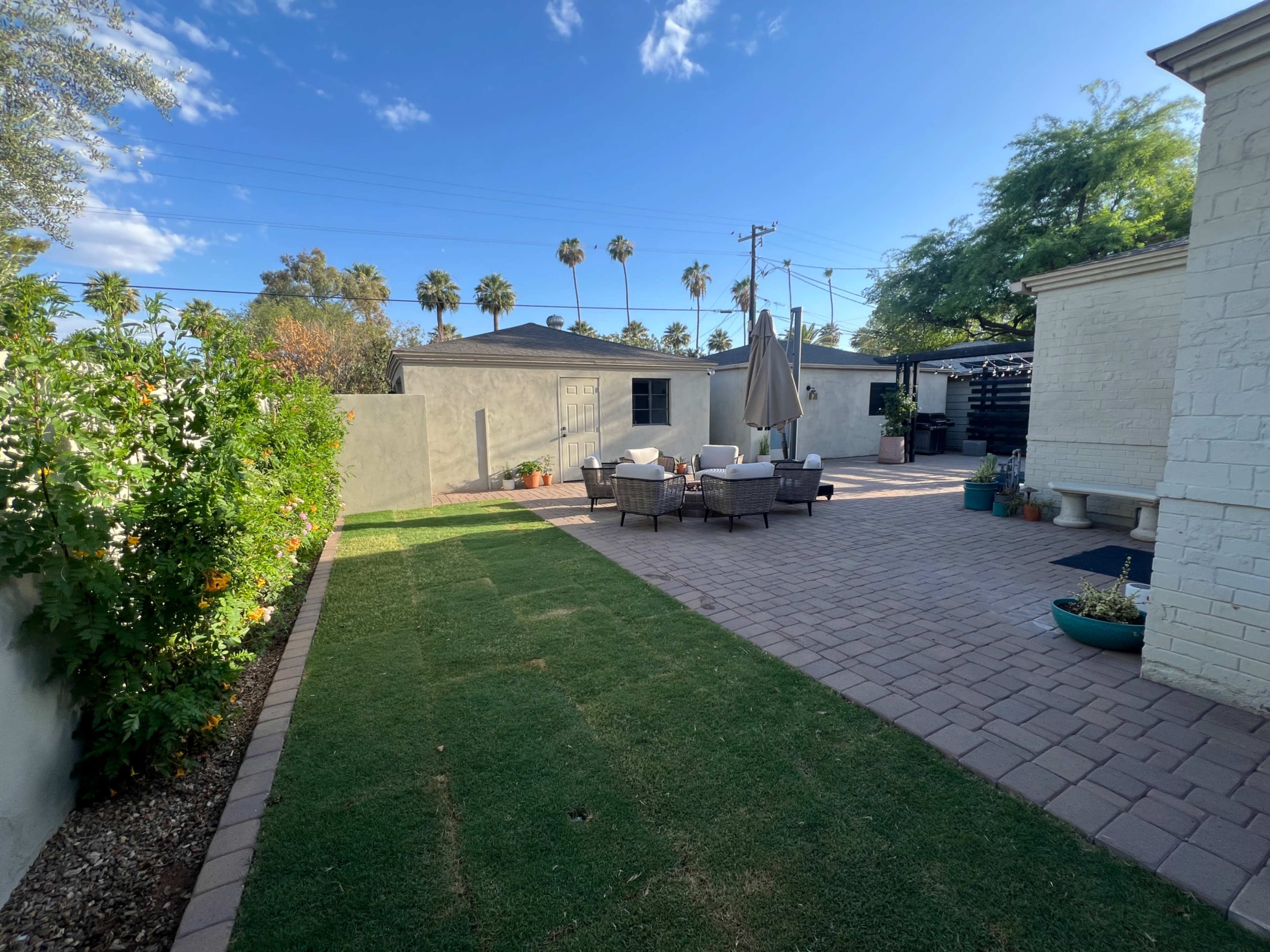 The image shows a backyard patio area with outdoor seating, a large umbrella, and a well-maintained lawn, bordered by walls and palm trees in the background.
