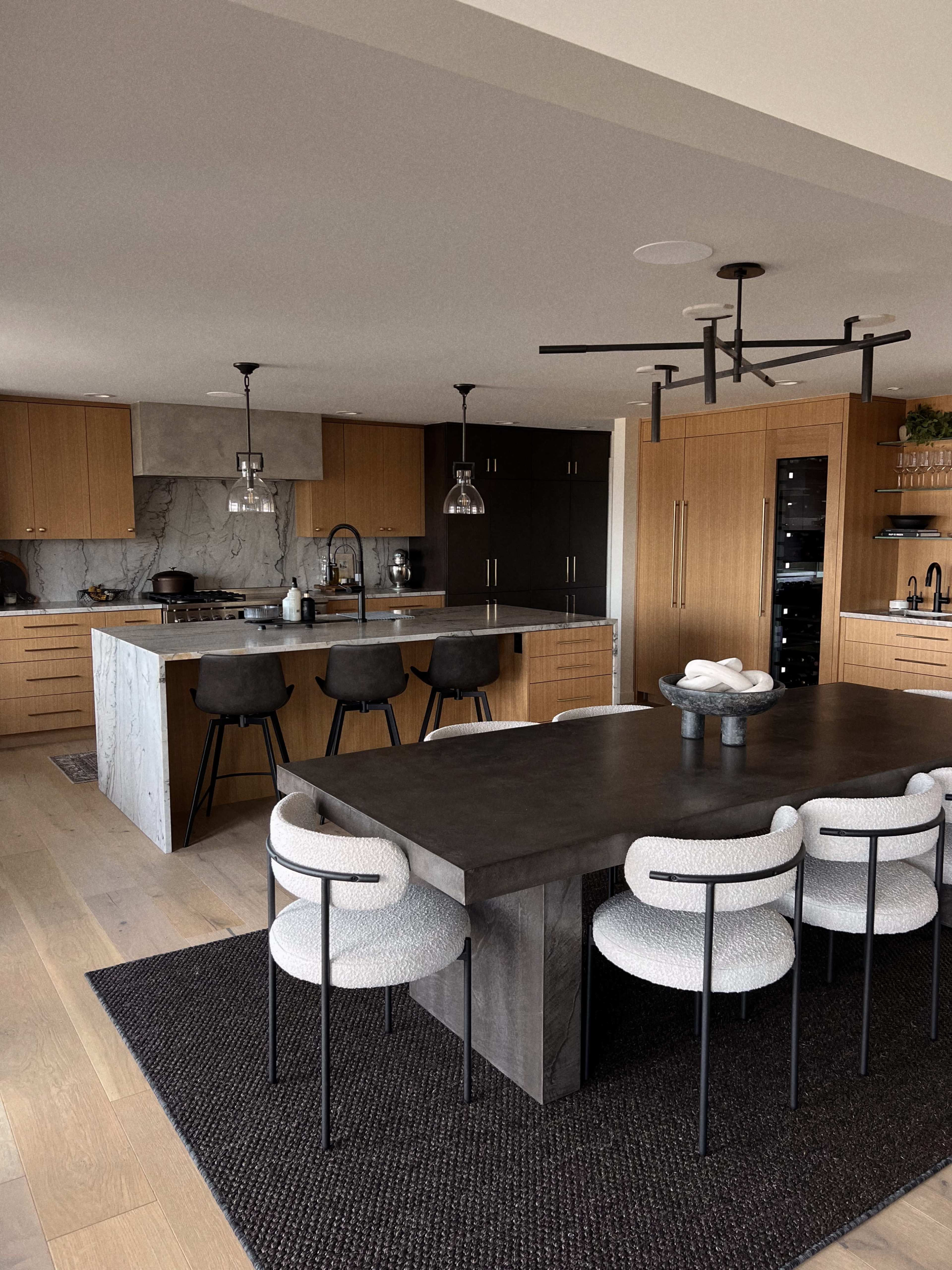 The image depicts a modern kitchen and dining area featuring a large dark table surrounded by upholstered chairs, with sleek cabinetry and pendant lighting above a kitchen island.