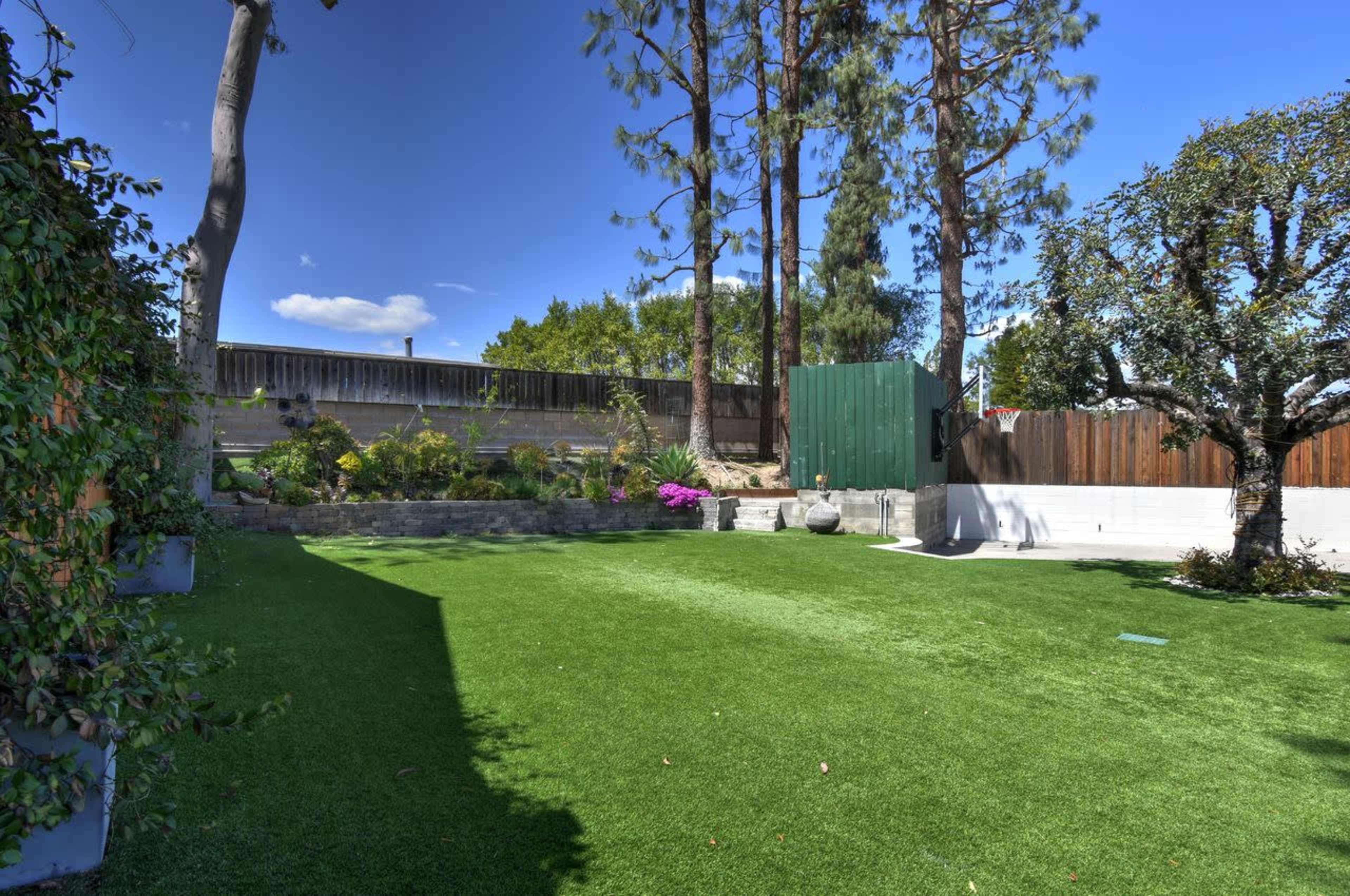 The image shows a landscaped backyard with artificial grass, surrounded by trees, a garden area, and a wooden fence.