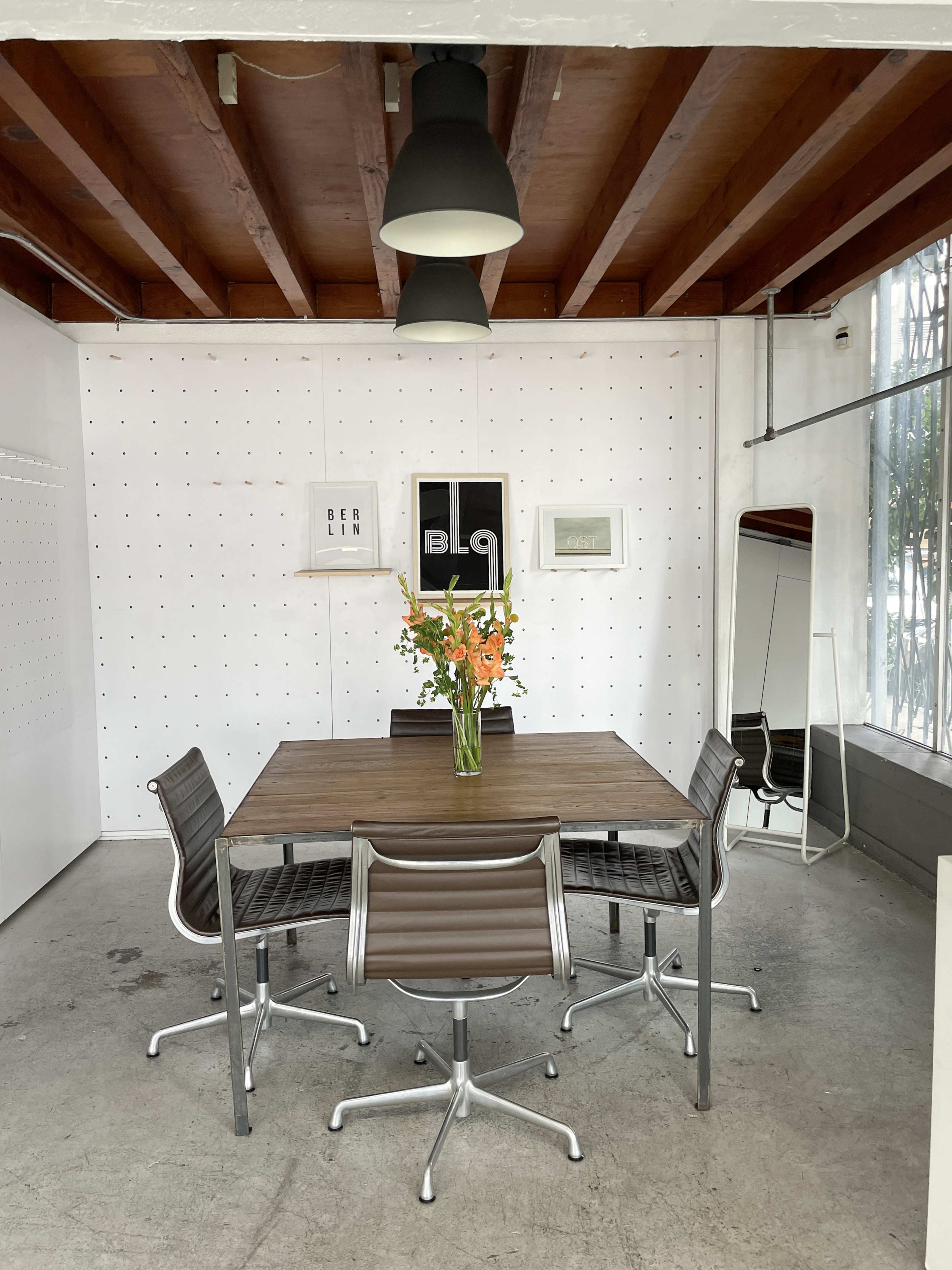 A wooden table surrounded by metal chairs is set in a bright room with white walls and overhead lighting, featuring a vase of flowers at the center.