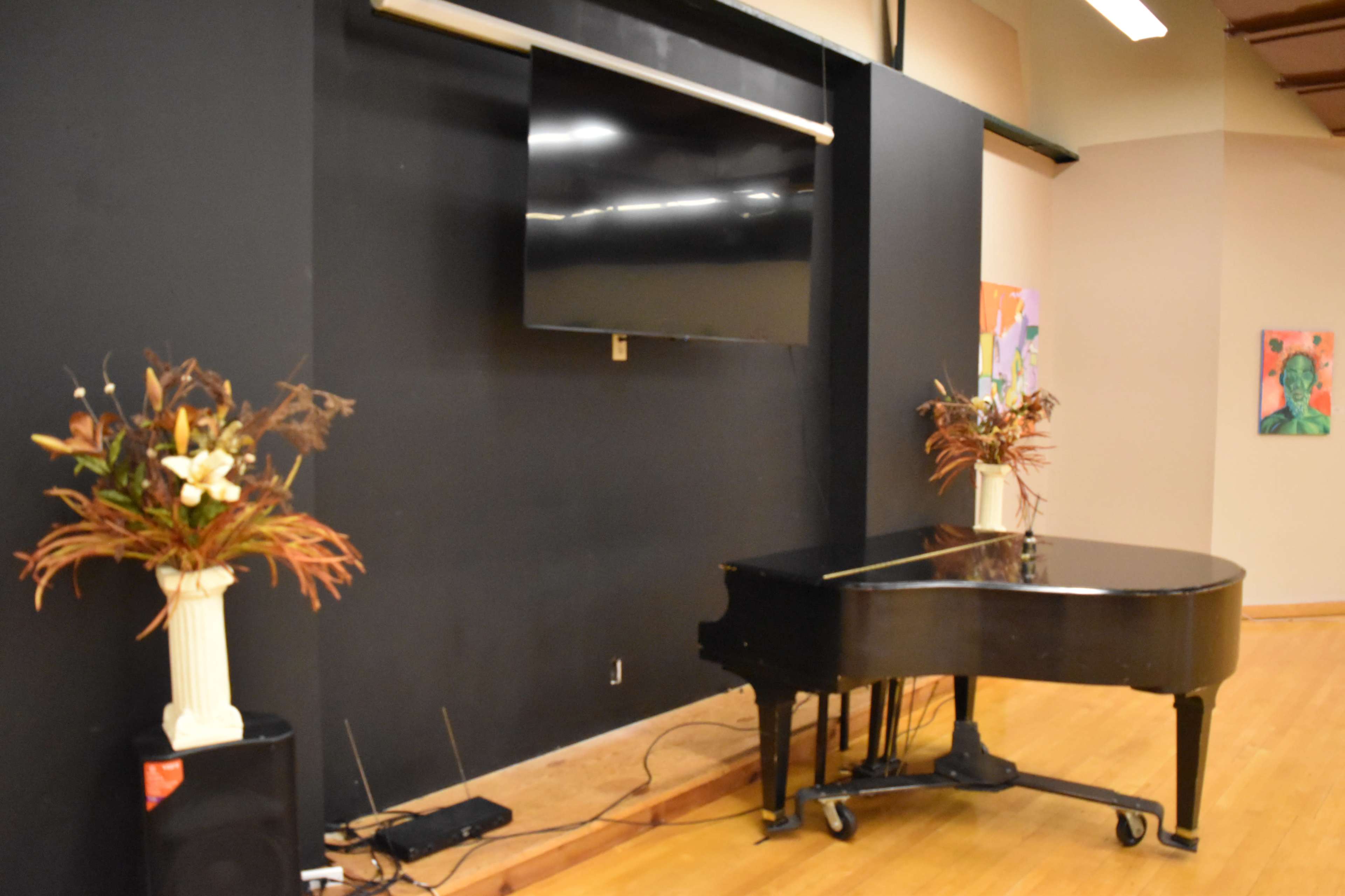 The image shows a black grand piano positioned in front of a wall with a mounted television, complemented by decorative floral arrangements on either side.