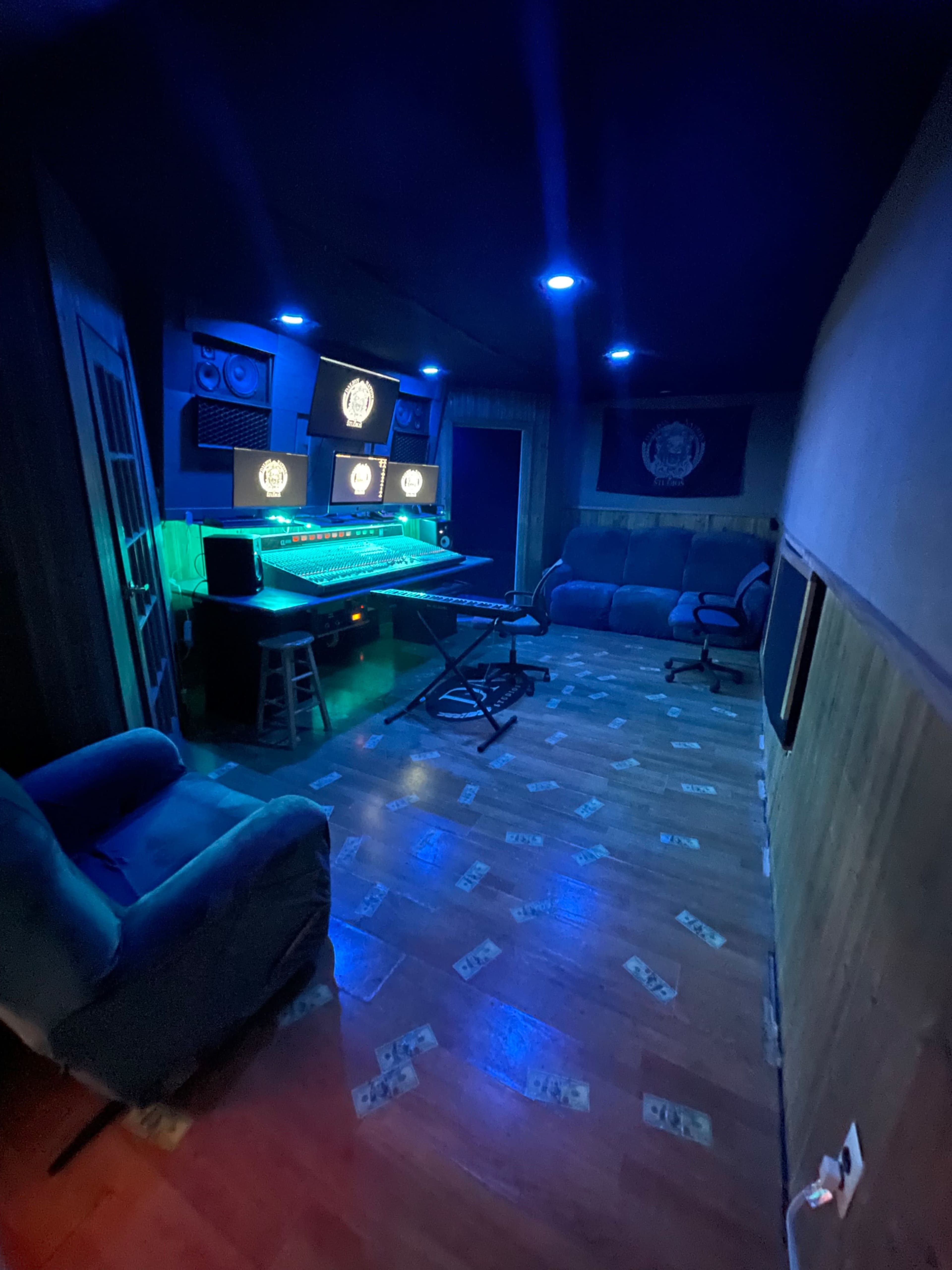 The image shows a dimly lit recording studio with a mixing board, multiple monitors, and blue lighting, featuring a seating area with two couches and a floor decorated with what appears to be money graphics.