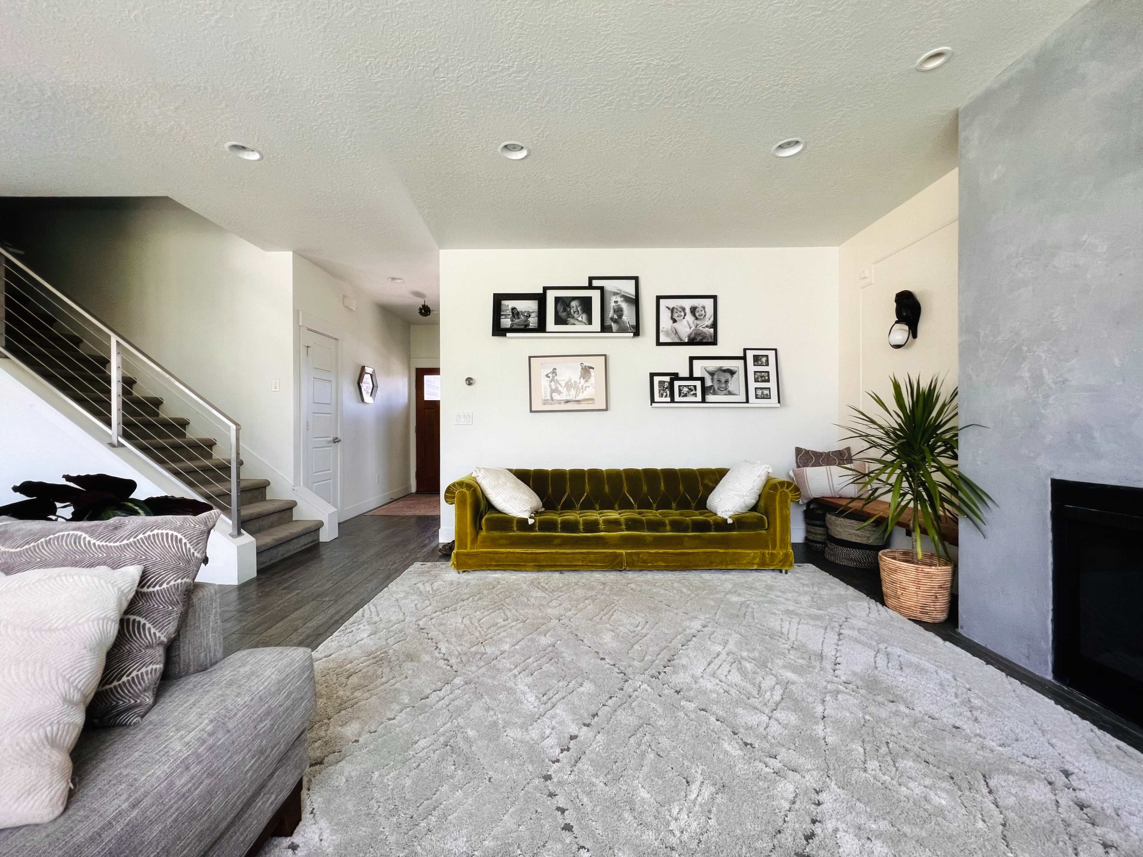 A modern living room features a yellow velvet couch, artwork on the walls, a staircase, and a large area rug.
